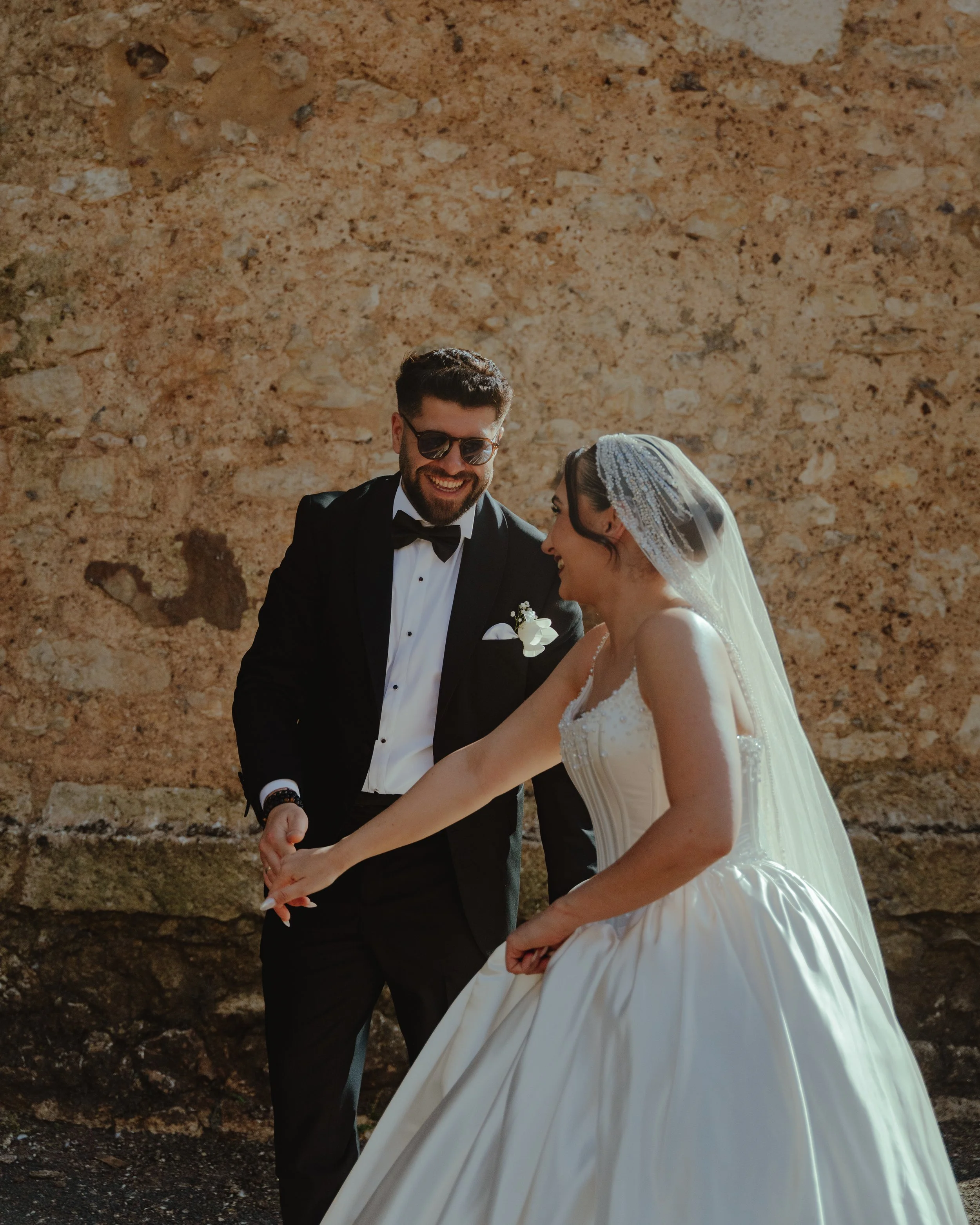 Un couple de mariés qui se tient la main, souriant et partageant un moment de bonheur devant un mur en pierre.