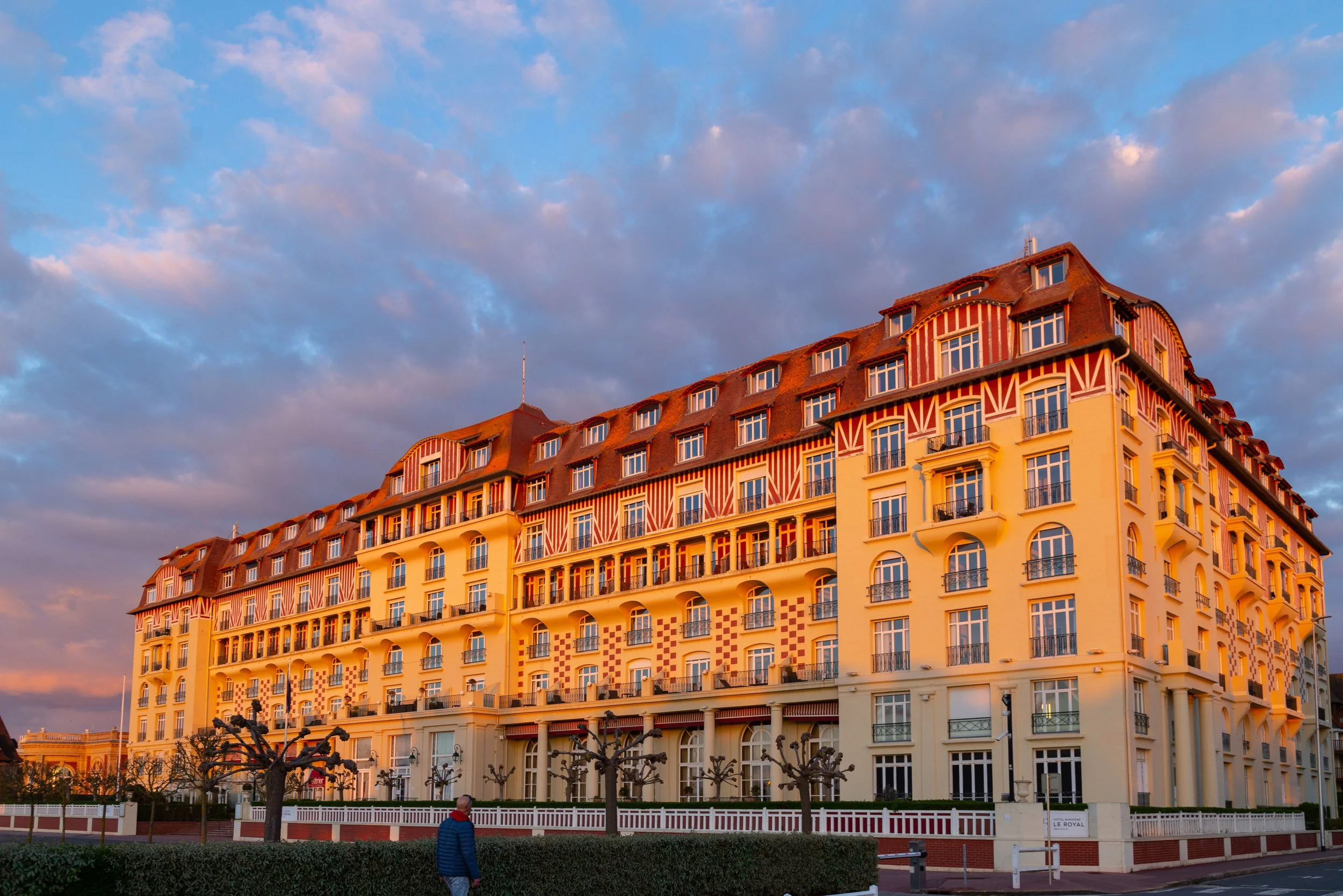 Un grand bâtiment résidentiel de style européen avec plusieurs étages, fenêtres et balcons, sous un ciel nuageux au coucher du soleil.