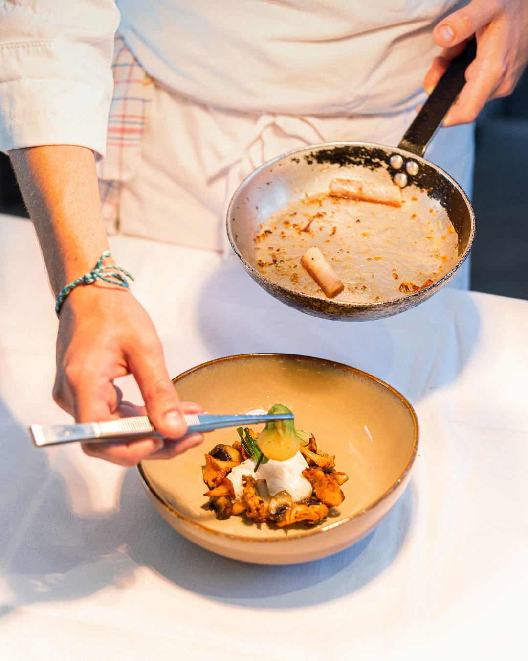 Une personne verse une sauce dans une assiette contenant des légumes et de la crème, sur une table blanche.