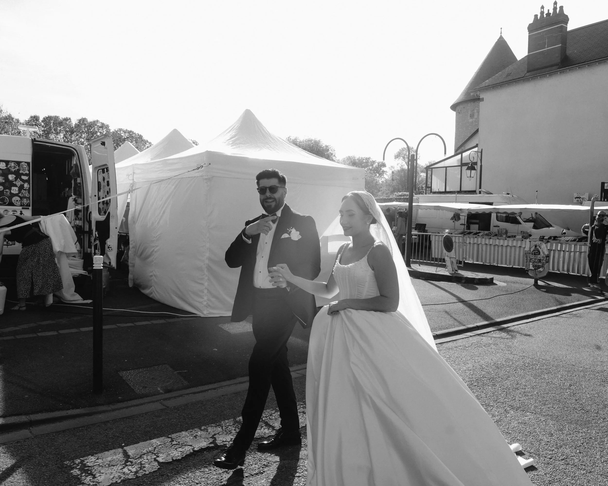 Un homme en costume de mariage et une femme en robe de mariage se tiennent devant des stands, avec un ciel clair en arrière-plan.
