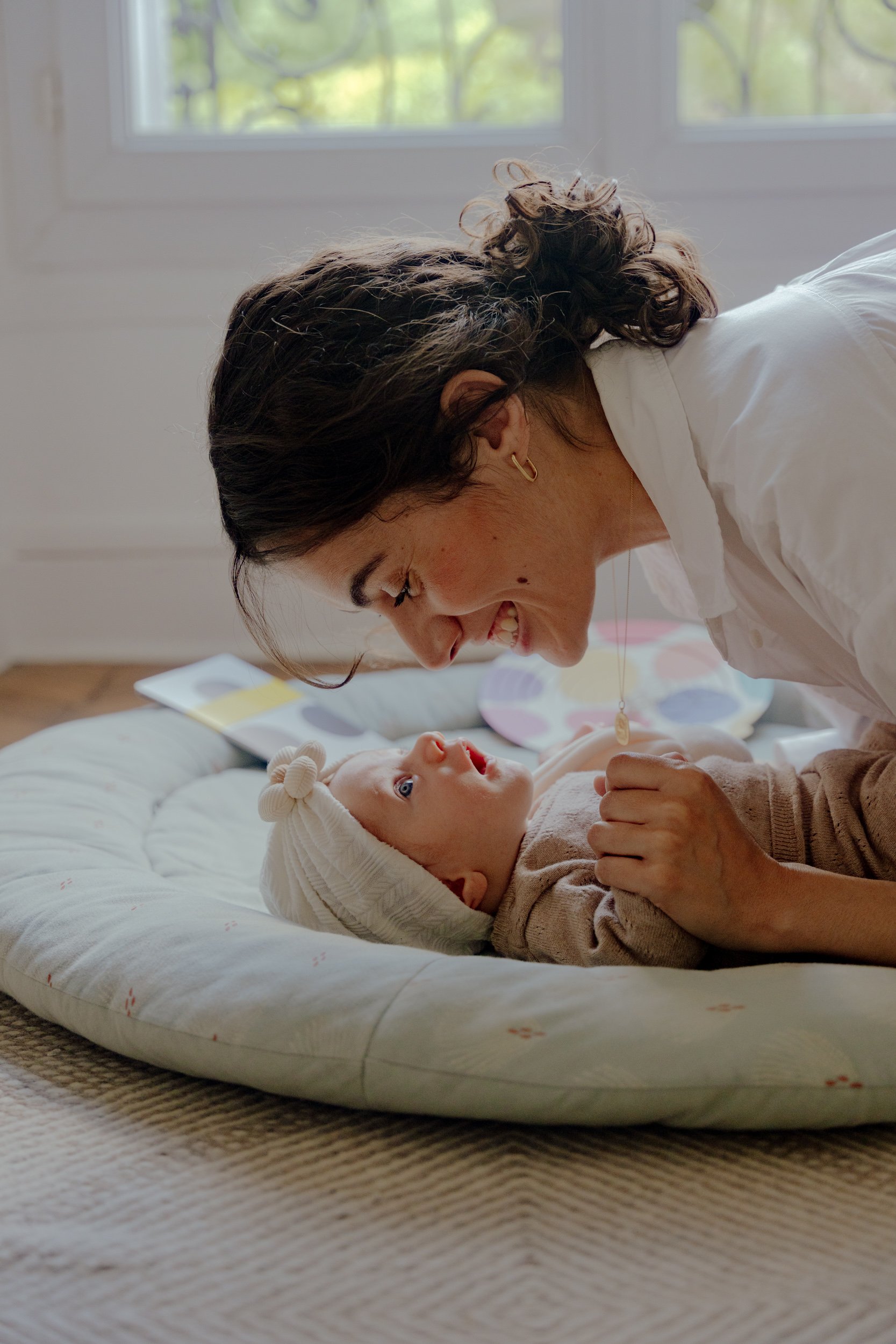 Une femme souriante se penche vers un bébé allongé sur un matelas, dans une pièce lumineuse avec une fenêtre en arrière-plan.