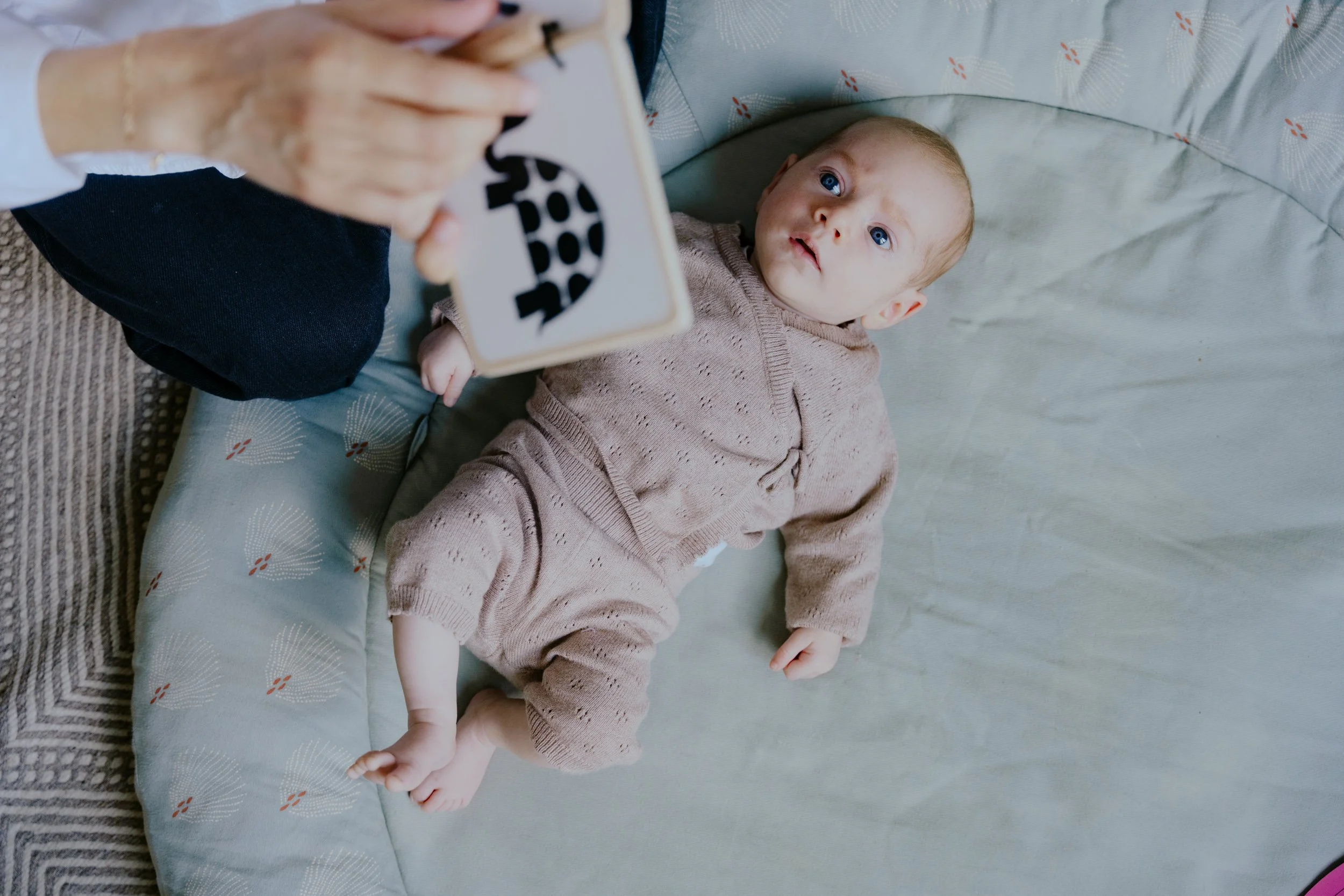 Un bébé allongé sur un lit regarde une personne qui lui montre un jouet en forme de lune avec des lettres. La personne tient le jouet devant le bébé, qui reste immobile. Le bébé porte un pull beige avec des petits trous.