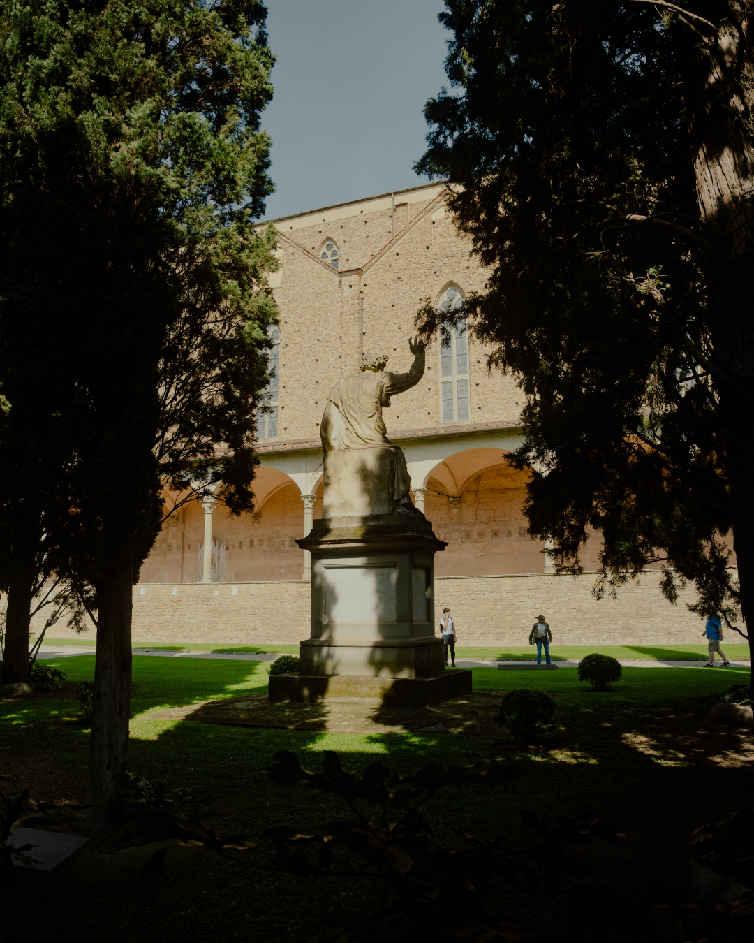Statue d'une figure humaine assise, vue de dos, avec un bras levé, devant un bâtiment en pierre avec des fenêtres gothiques, entourée d'arbres et de jeunes personnes dans un parc.