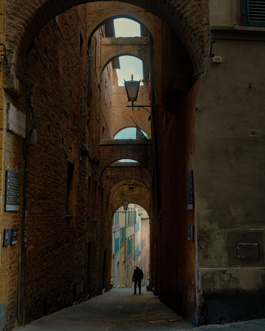 Une ruelle étroite avec des arches en briques, des lampadaires anciens, et une silhouette humaine marchant au loin.