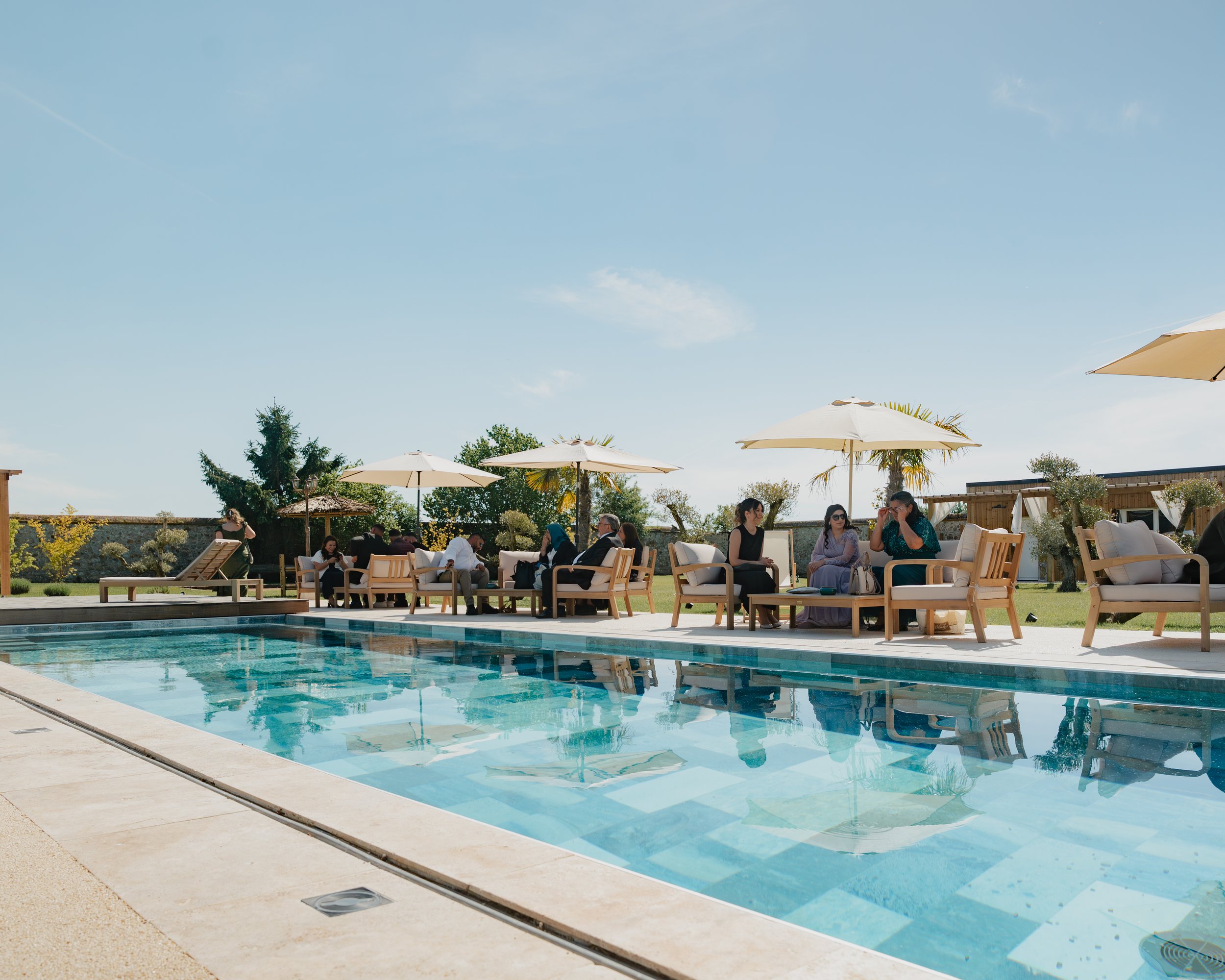 Piscine extérieure avec des chaises longues en bois, des parasols et des gens assis ou se relaxant sous un ciel ensoleillé.
