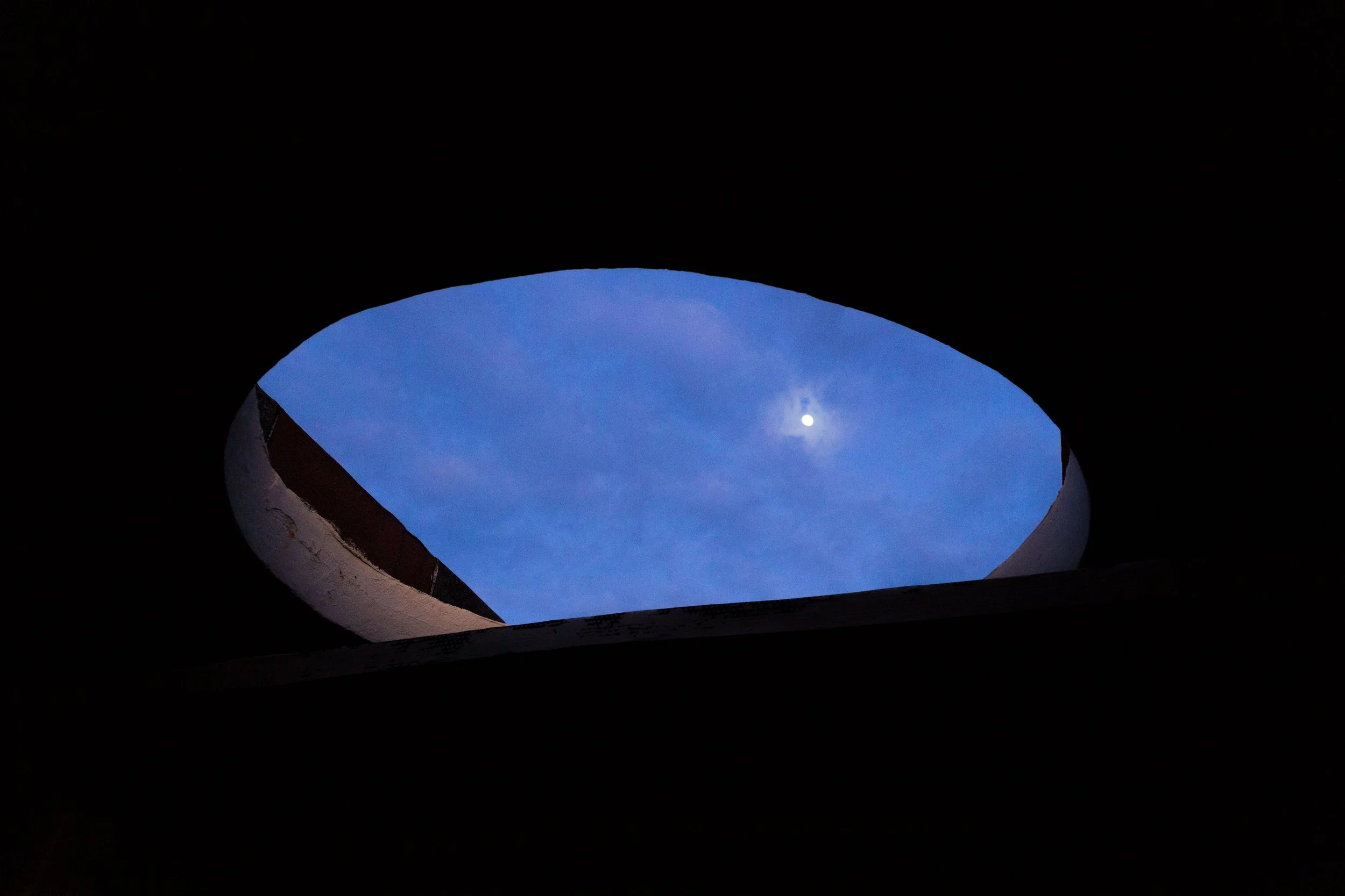 Ciel nocturne vu à travers un trou arrondi dans un plafond ou un mur, avec la lune visible dans le ciel bleu clair.