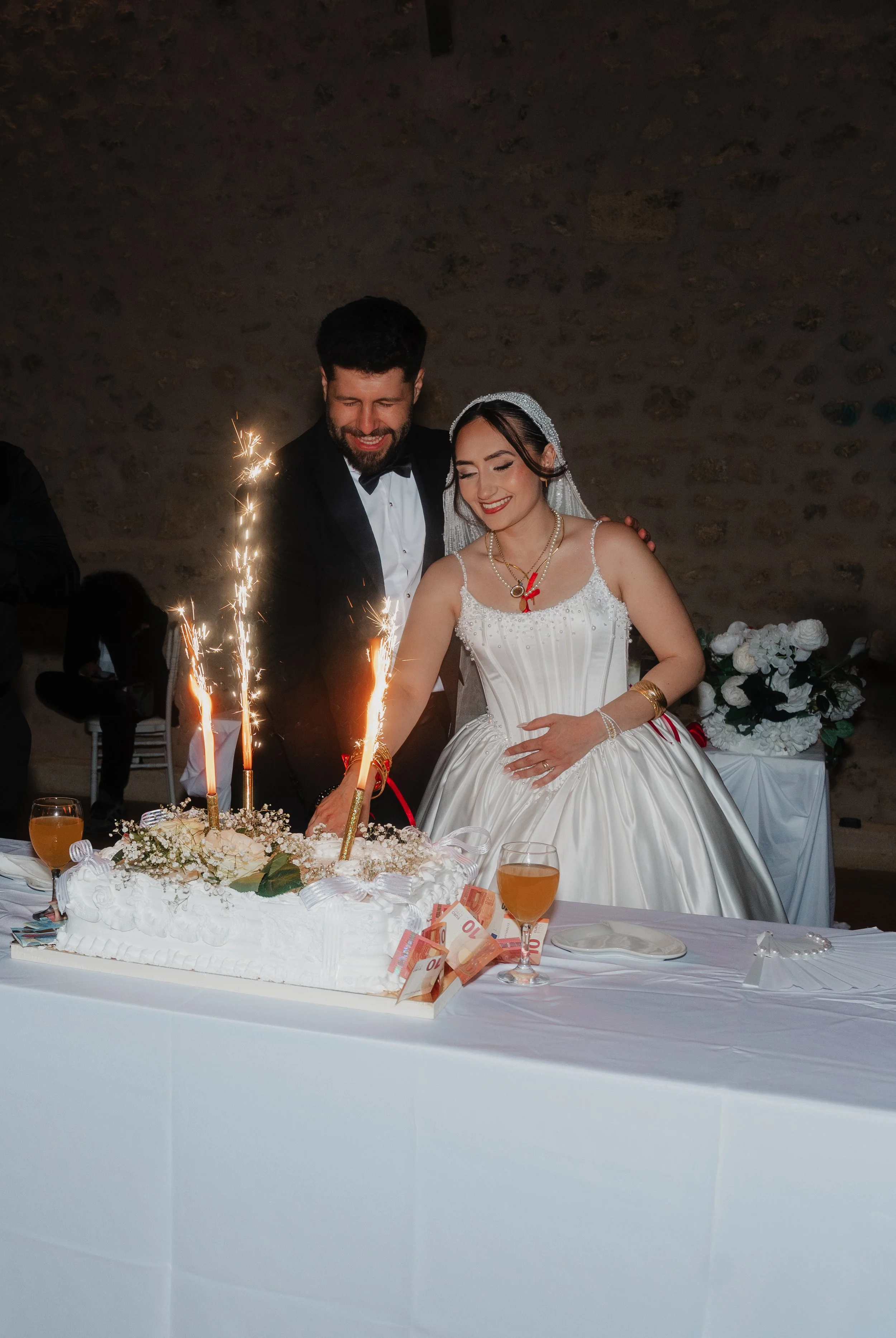 Un couple de mariés coupe un gâteau de mariage décoré de fleurs blanches, avec des bougies et des étincelles, lors d'une célébration de mariage dans une salle avec un mur en pierre.