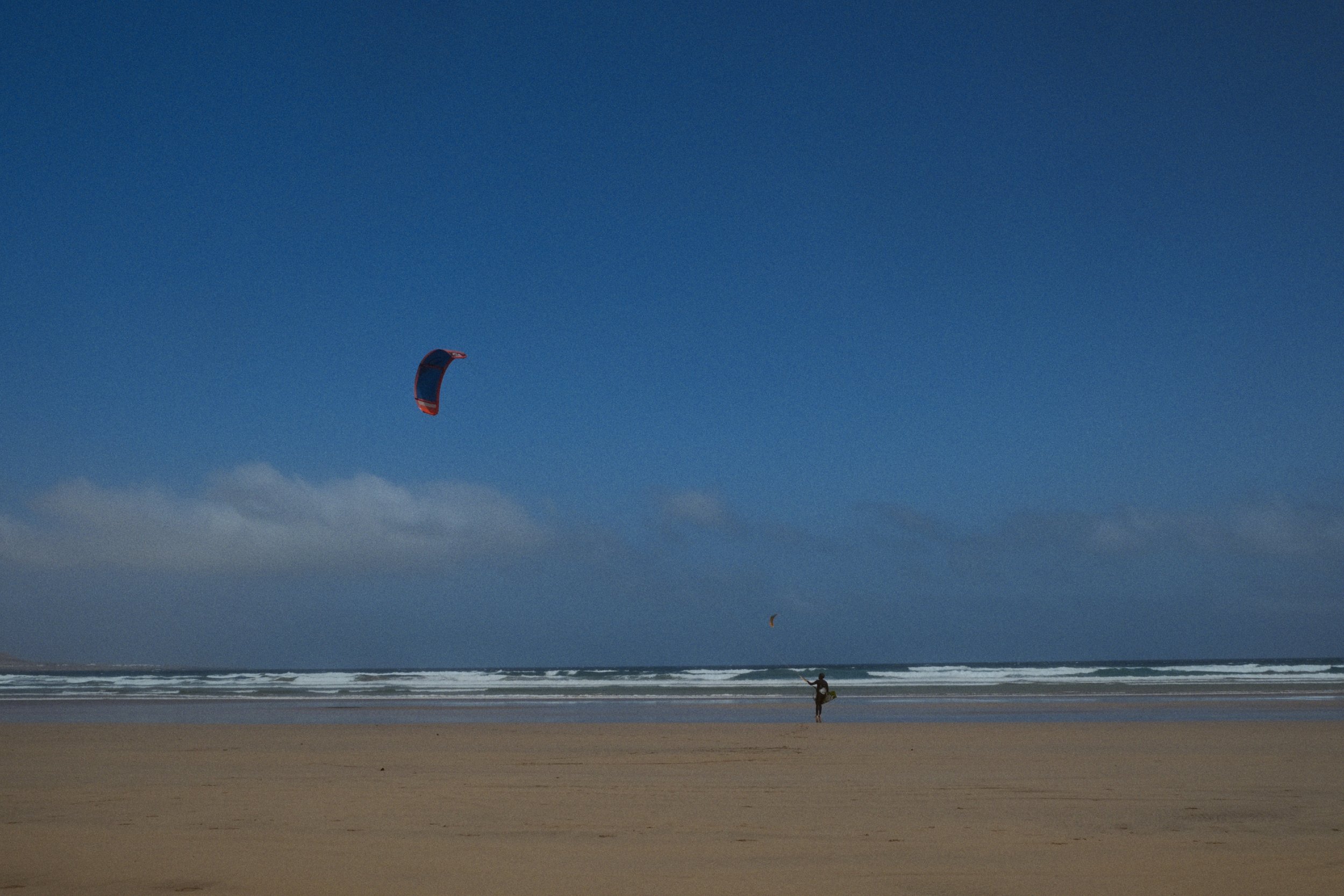 Un rider de kitesurf navigue sur la mer avec le cerf-volant au sommet de sa trajectoire dans un ciel bleu clair, près d'une plage de sable vidée, avec une petite silhouette de personne en bas. La scène est saisissante et évoque la liberté et l'aventu