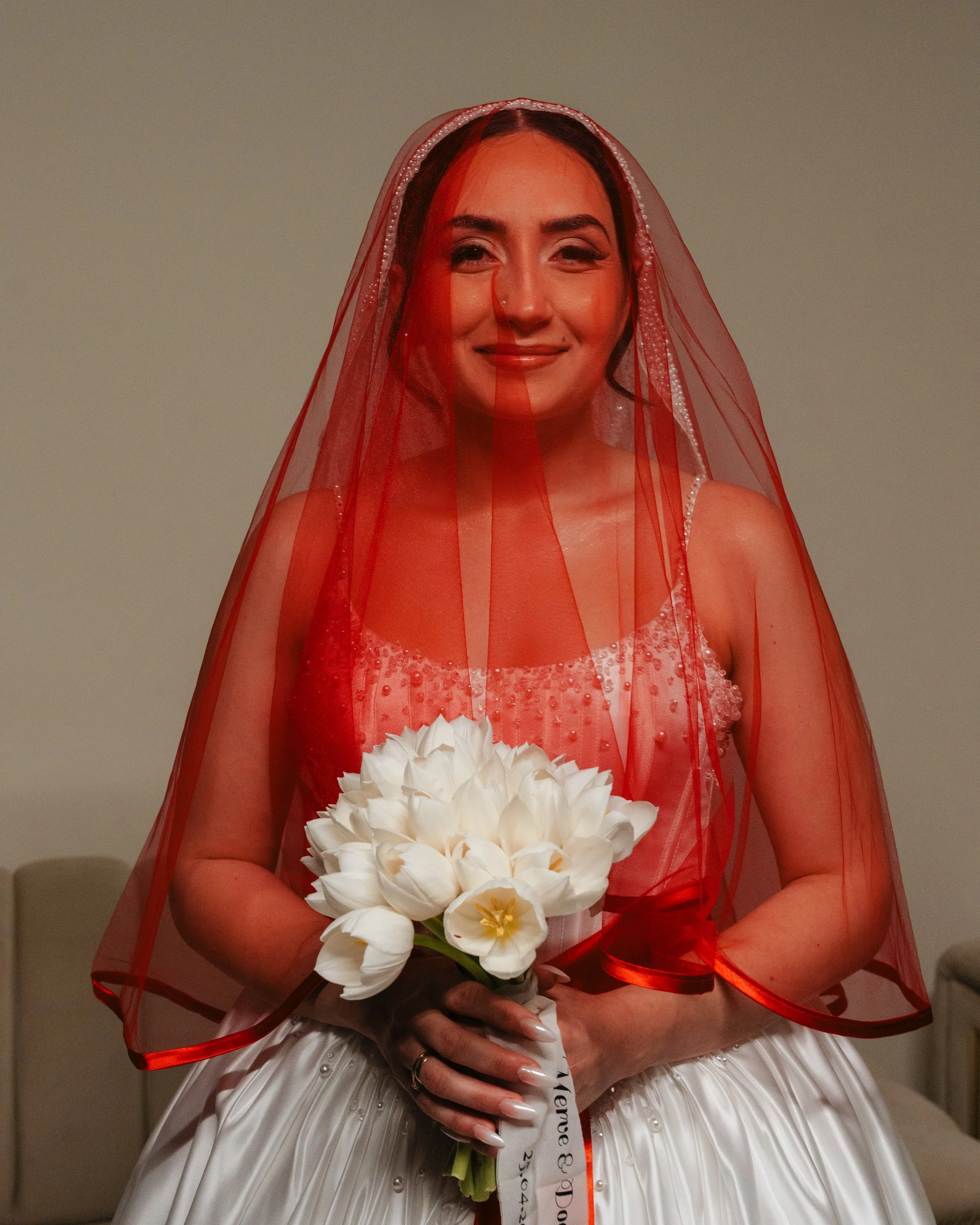 Une femme portant une robe blanche avec des détails perlés, coiffée d'une voilette rouge avec des bords en dentelle, tenant un bouquet de fleurs blanches. La femme sourit doucement.