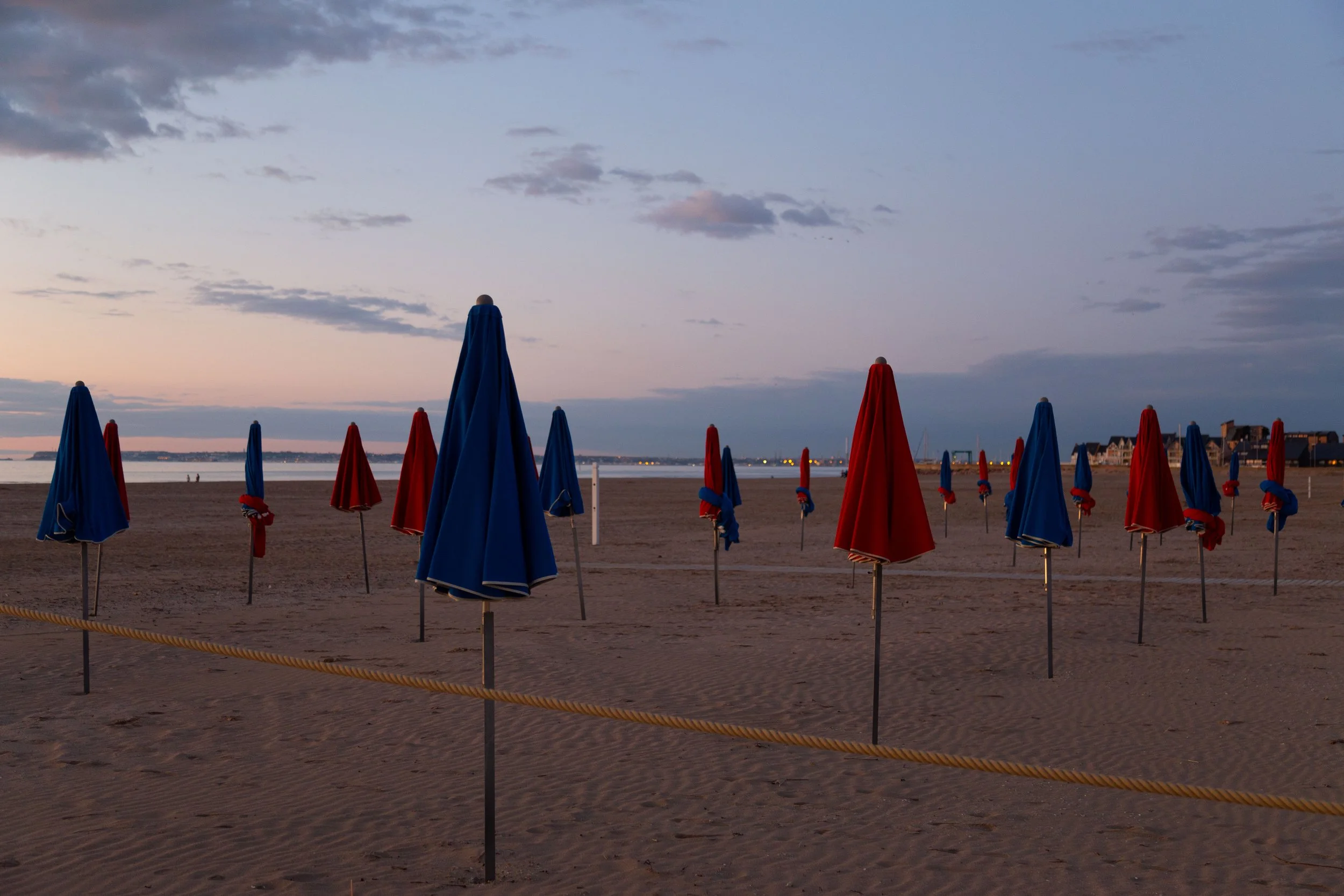 Parasols bleus et rouges fermés au bord de la plage au coucher du soleil, avec un ciel partiellement nuageux et une ville en arrière-plan.