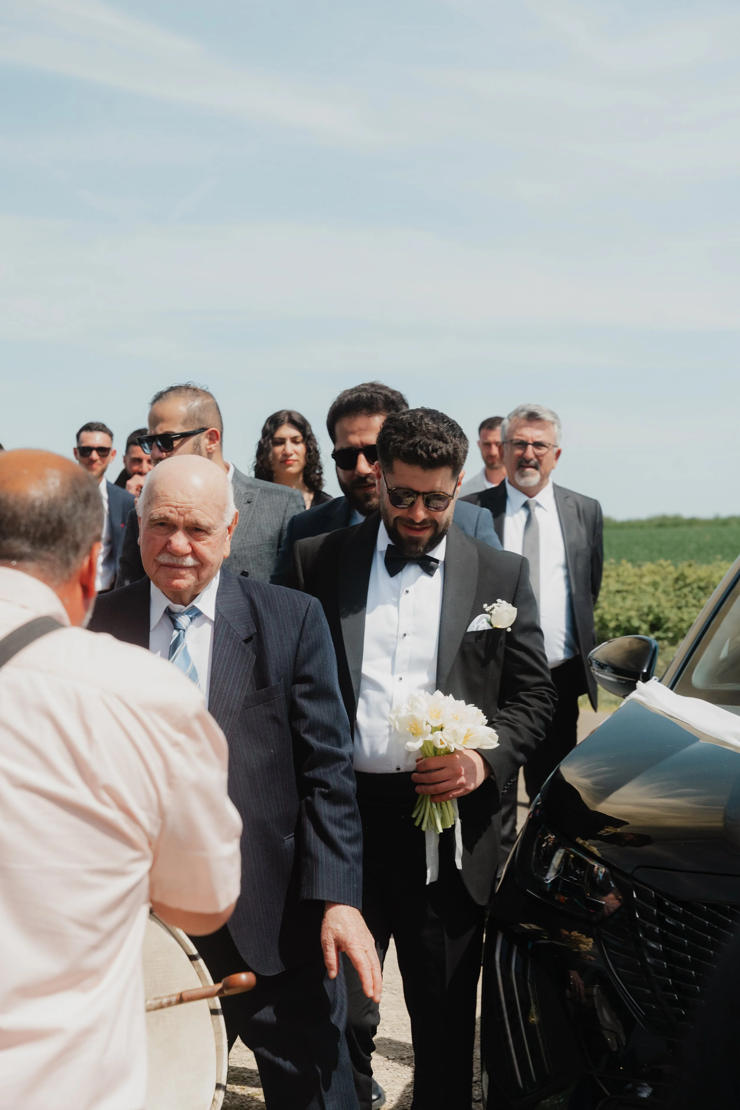 Groupe de personnes en costume lors d'une cérémonie en plein air, avec un homme en costume noir tenant un bouquet de fleurs blanches, en face d'une voiture noire.