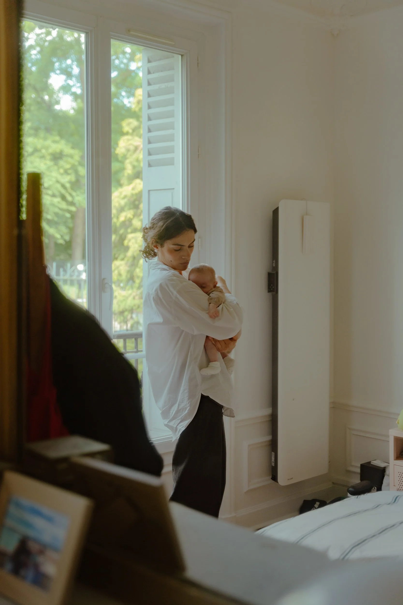 Une femme tenant un bébé dans une pièce lumineuse près d'une fenêtre ouverte, vue partielle depuis une porte ou un miroir.