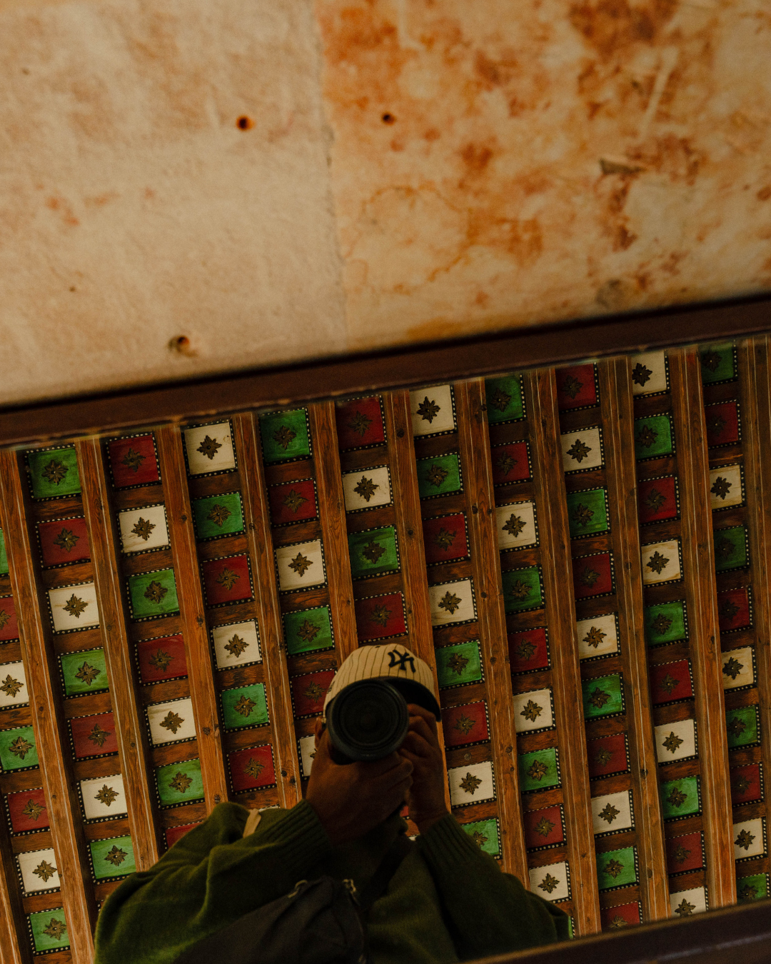 Reflet d'une personne prenant une photo dans un miroir, avec un plafond en bois décoré de carreaux colorés en arrière-plan.