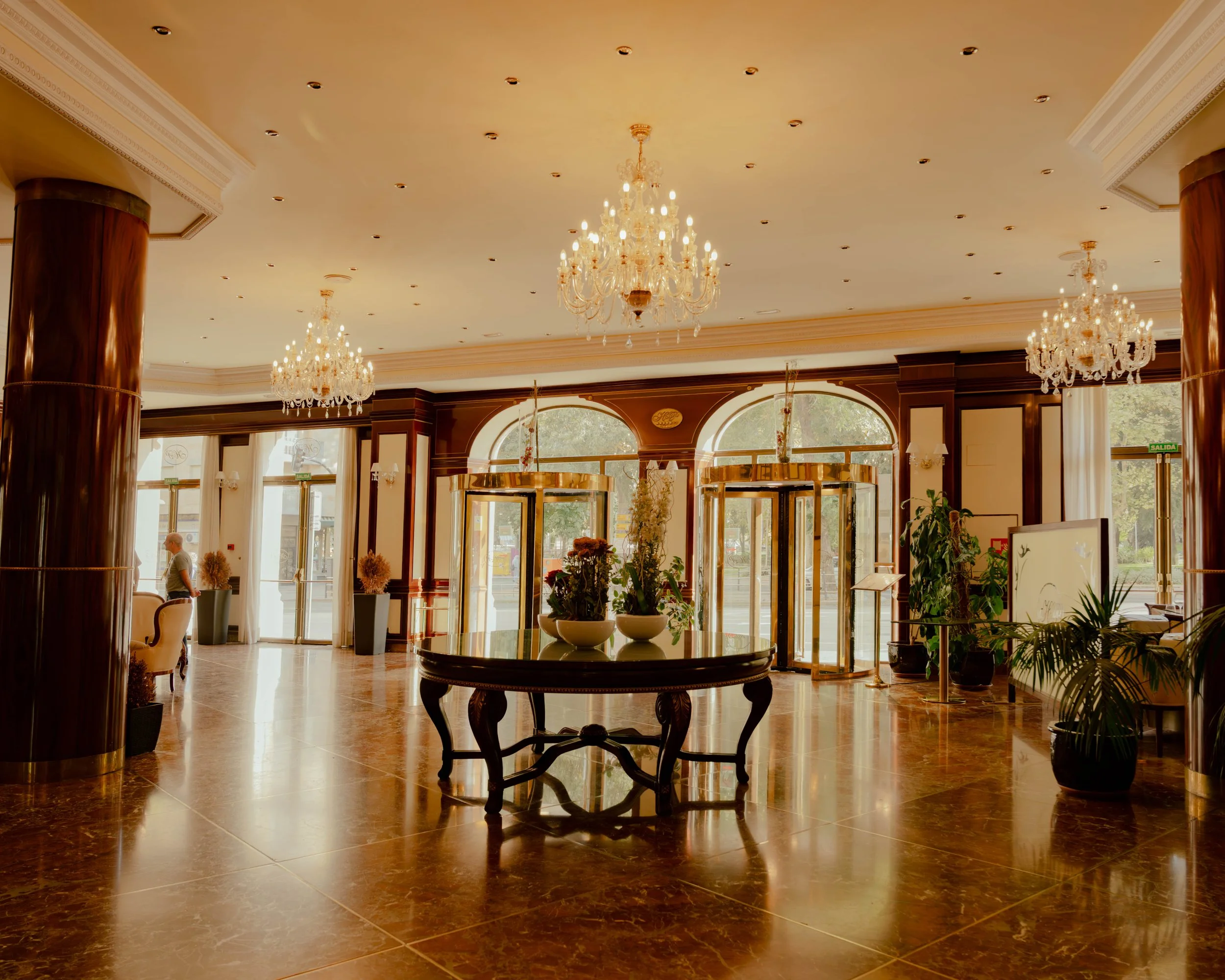 Intérieur d'un hall de réception avec un plafond orné de lustres en cristal, grande table ronde en bois, colonnes en bois laqué, grande entrée avec portes en verre, plantes en pot, et un person en arrière-plan.