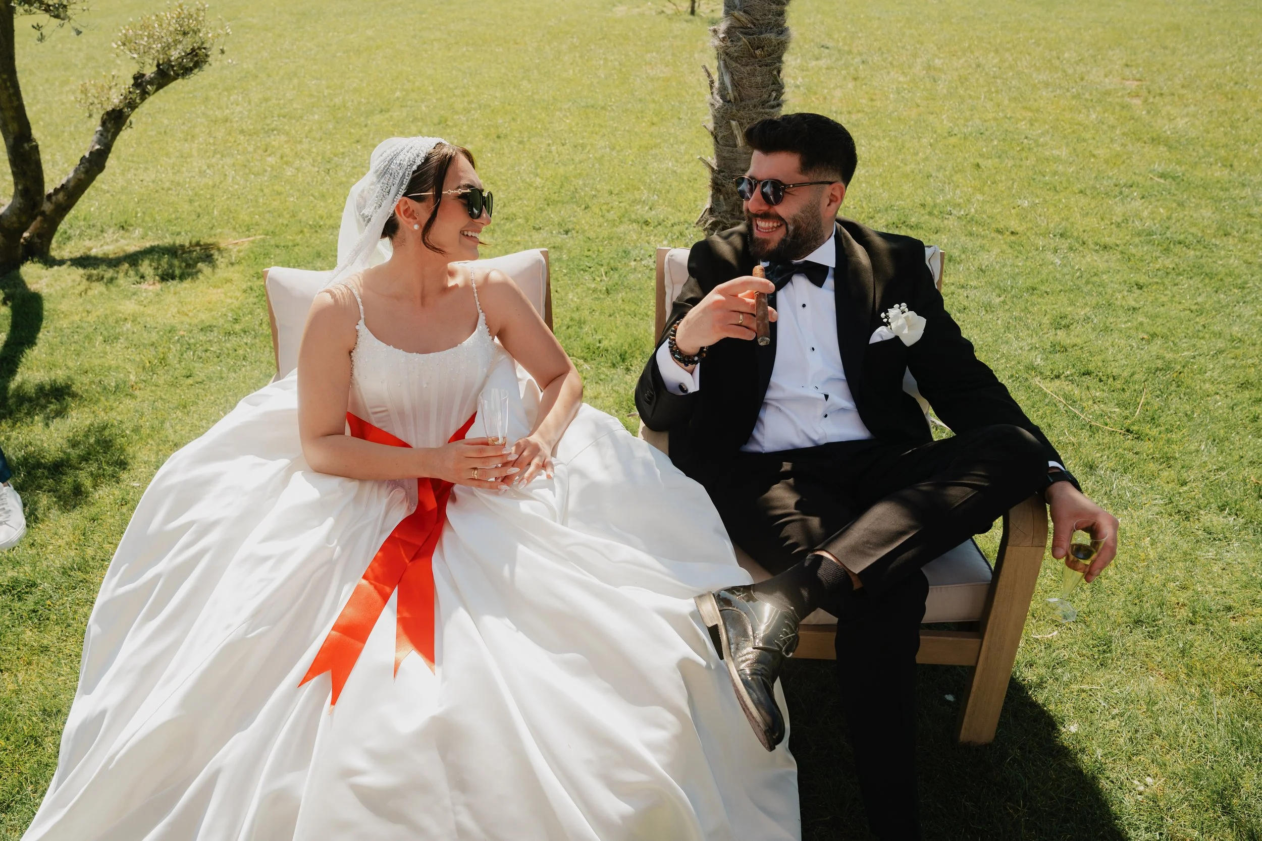 Un couple souriant en tenue de mariage, assis dans un jardin ensoleillé, partageant un moment joyeux avec une cigare et des boissons.