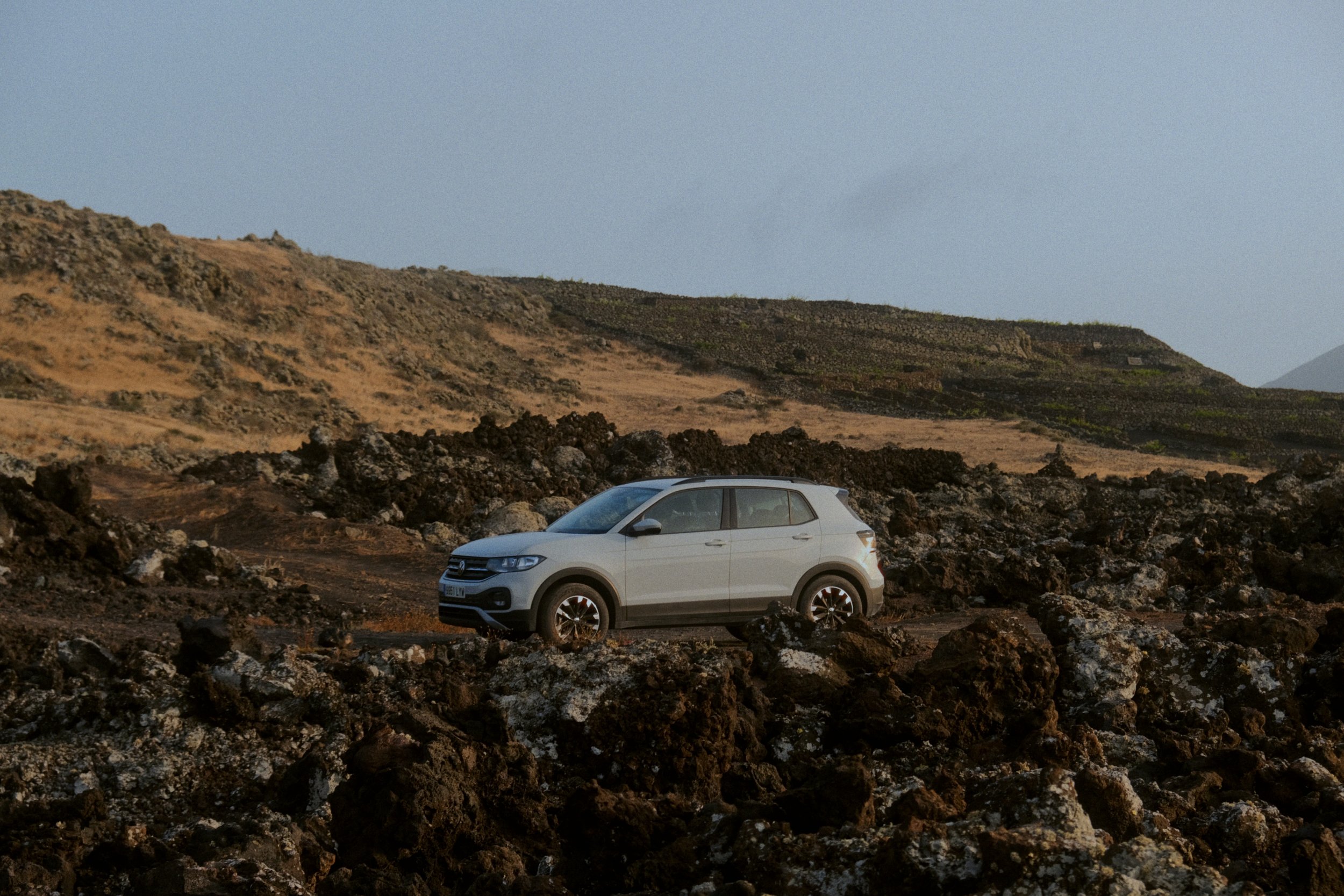 Une voiture blanche SUV sur un terrain rocheux dans un paysage de montagnes avec des collines en arrière-plan, sous un ciel clair.
