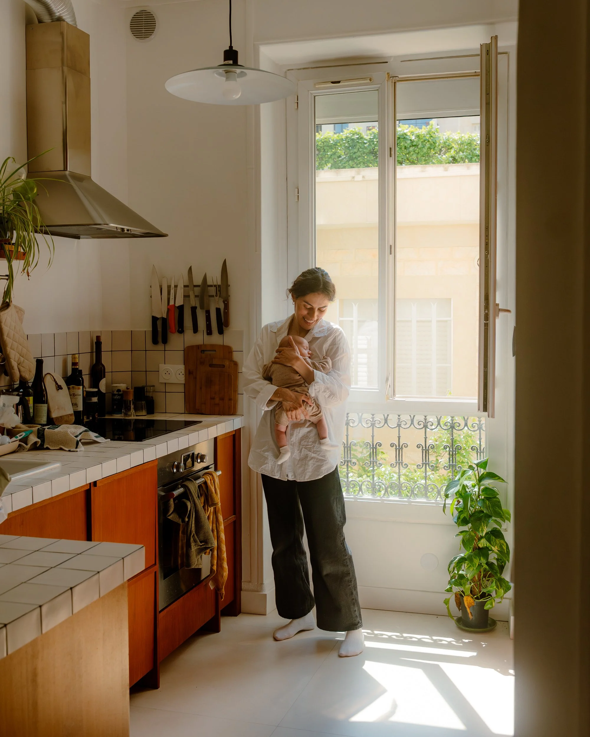 Une femme souriante tient un bébé dans la cuisine près d'une fenêtre ouverte avec un balcon. La pièce est lumineuse, il y a des ustensiles de cuisine et des bouteilles sur le plan de travail, et une plante en pot est visible à côté de la fenêtre.
