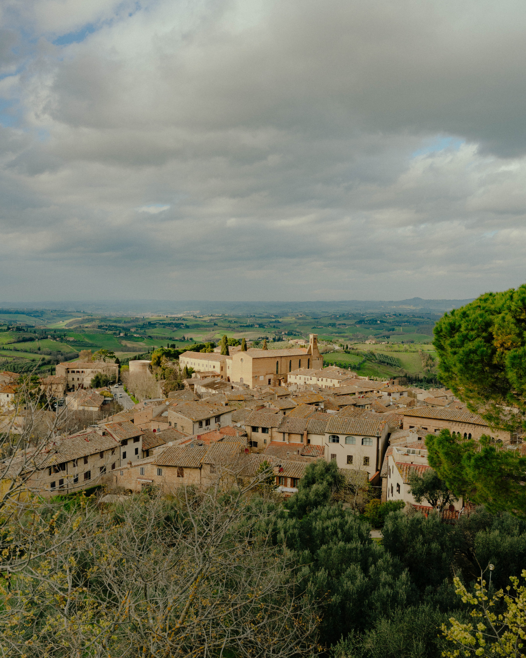 Vue panoramique d'un village médiéval avec bâtiments en pierre et un grand édifice religieux, entouré de collines verdoyantes sous un ciel nuageux.