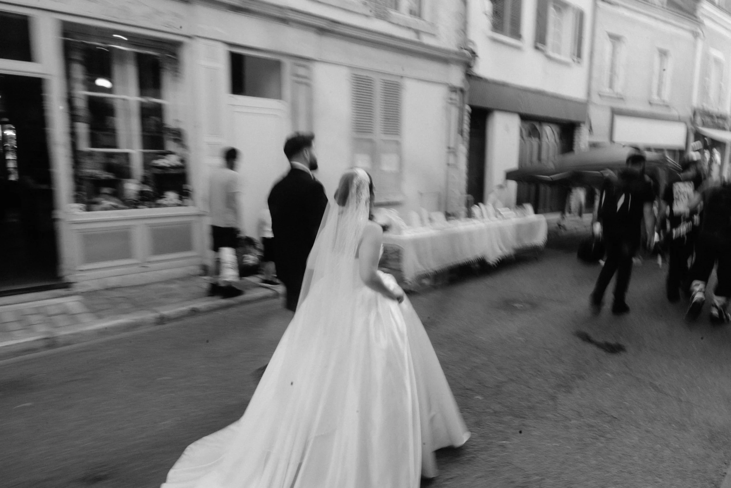 Une femme en robe de mariage blanche marche dans la rue, accompagnée d'autres personnes, dans un environnement urbain avec des bâtiments en arrière-plan.