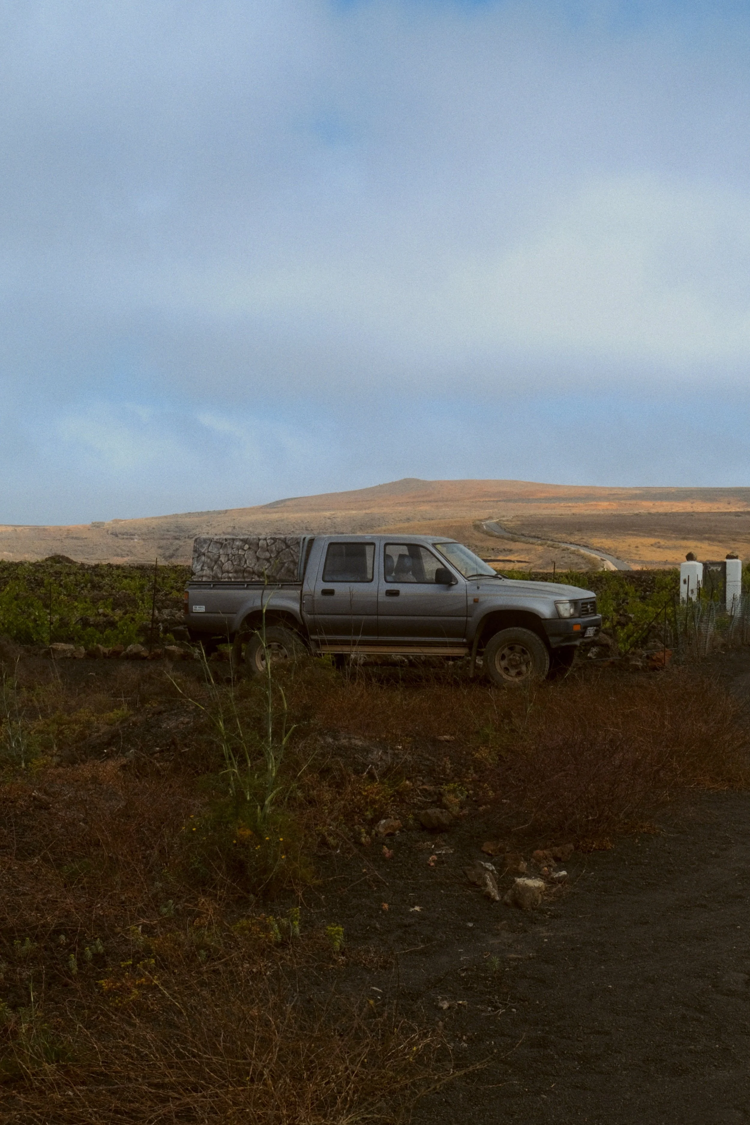 Une voiture grise stationnée dans un paysage désertique avec des collines en arrière-plan et un ciel partiellement nuageux.