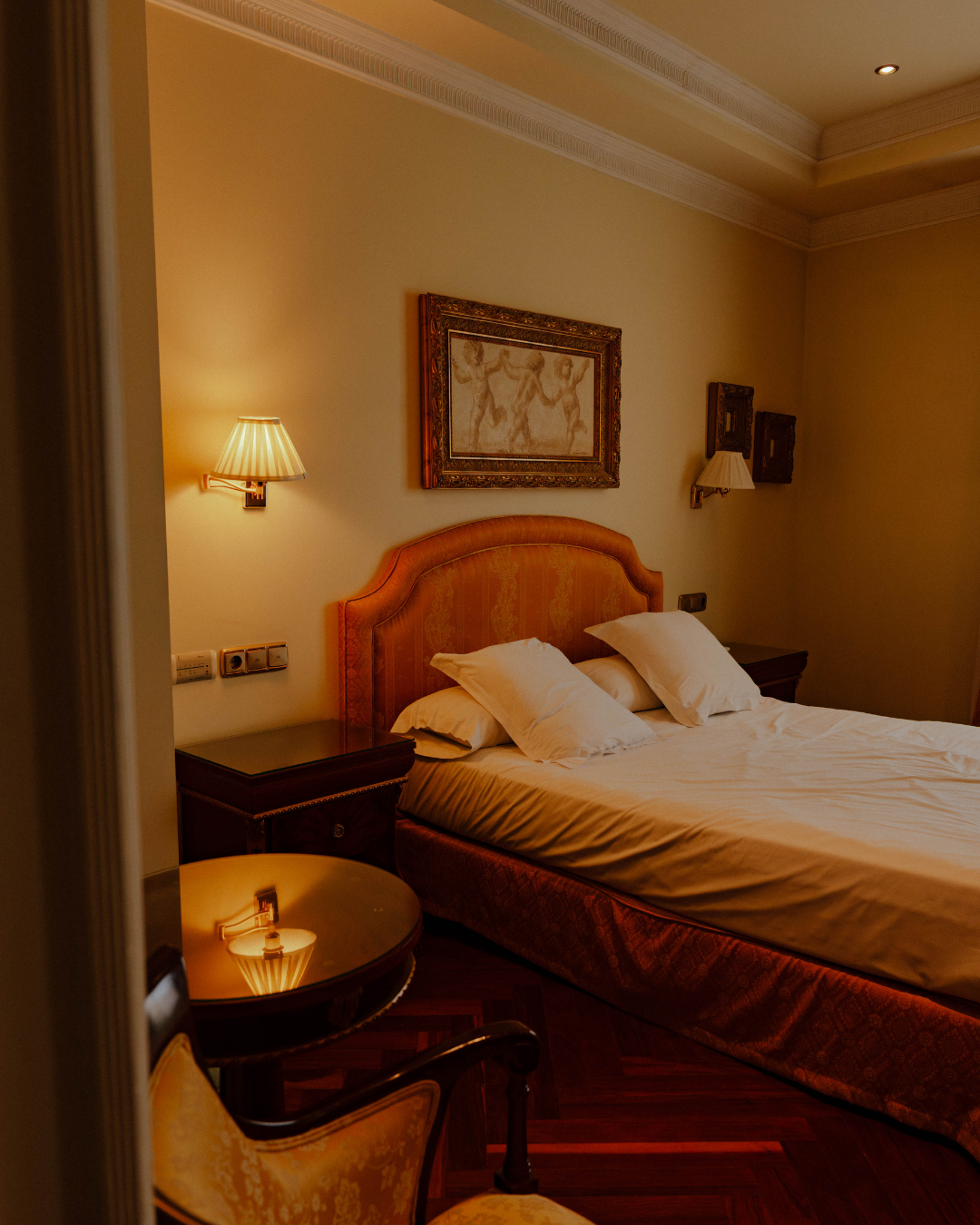 Chambre d'hôtel avec lit king-size, tête de lit en tissu orange, deux lampes murales, tableau au-dessus du lit, table ronde en verre, fauteuil en bois avec coussin, murs colorés, plafond avec moulures.