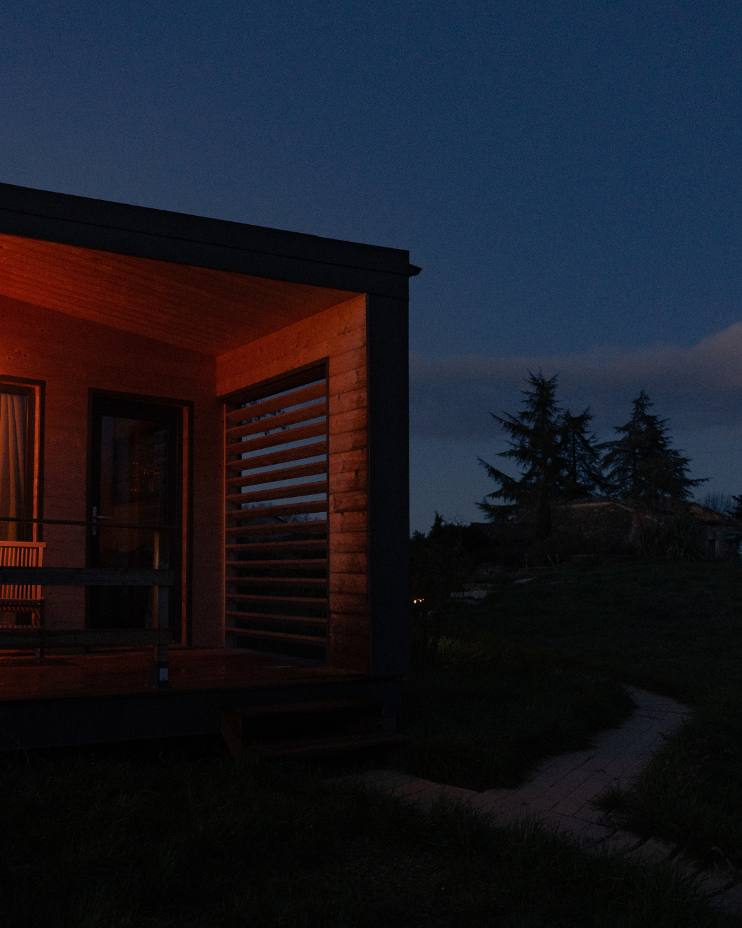 Une maison moderne en bois la nuit, avec une lumière chaude à l'intérieur, entourée d'arbres et un ciel sombre.