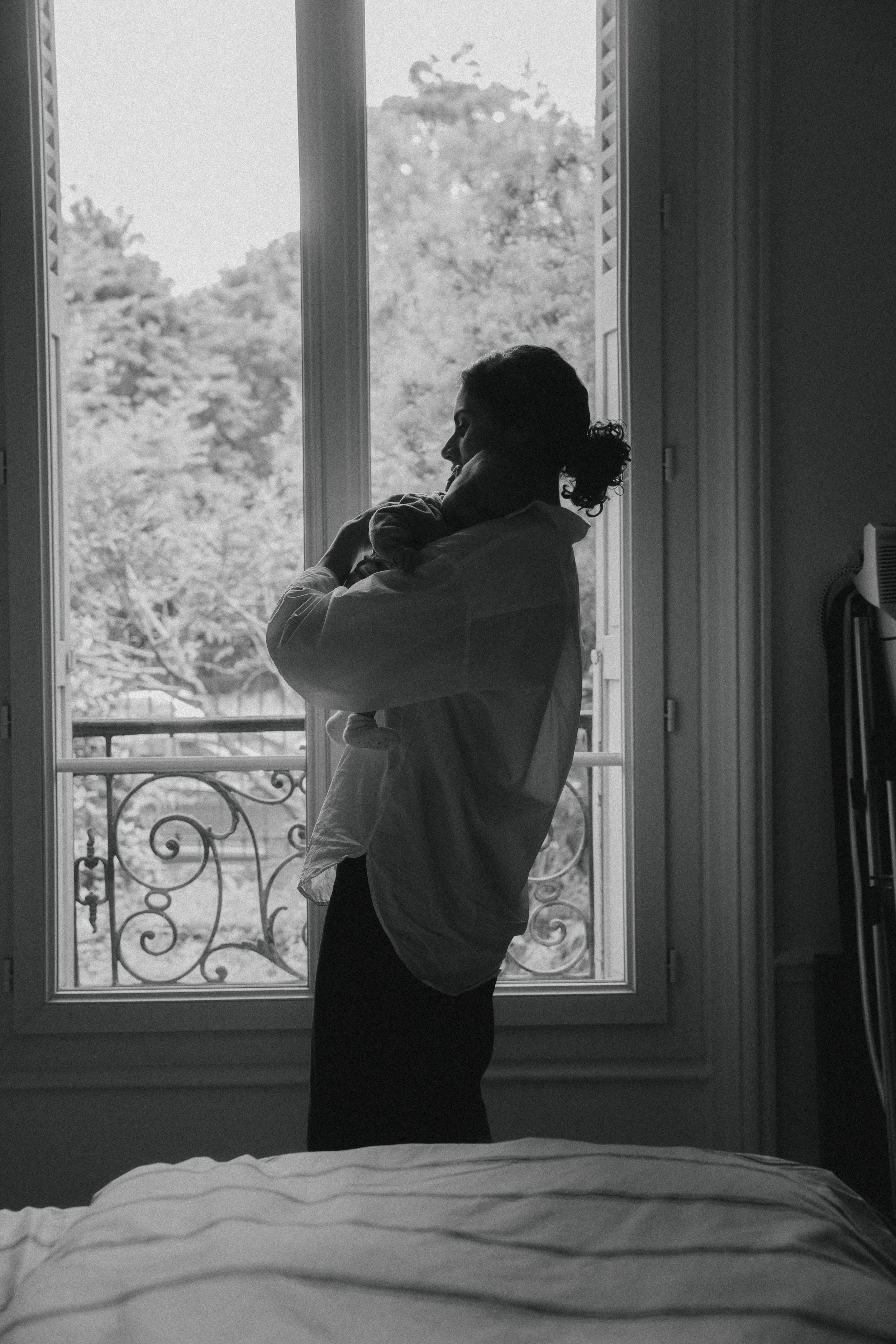 Une personne debout près d'une grande fenêtre, tenant un enfant dans ses bras, dans une pièce sombre avec une vue sur des arbres à l'extérieur.