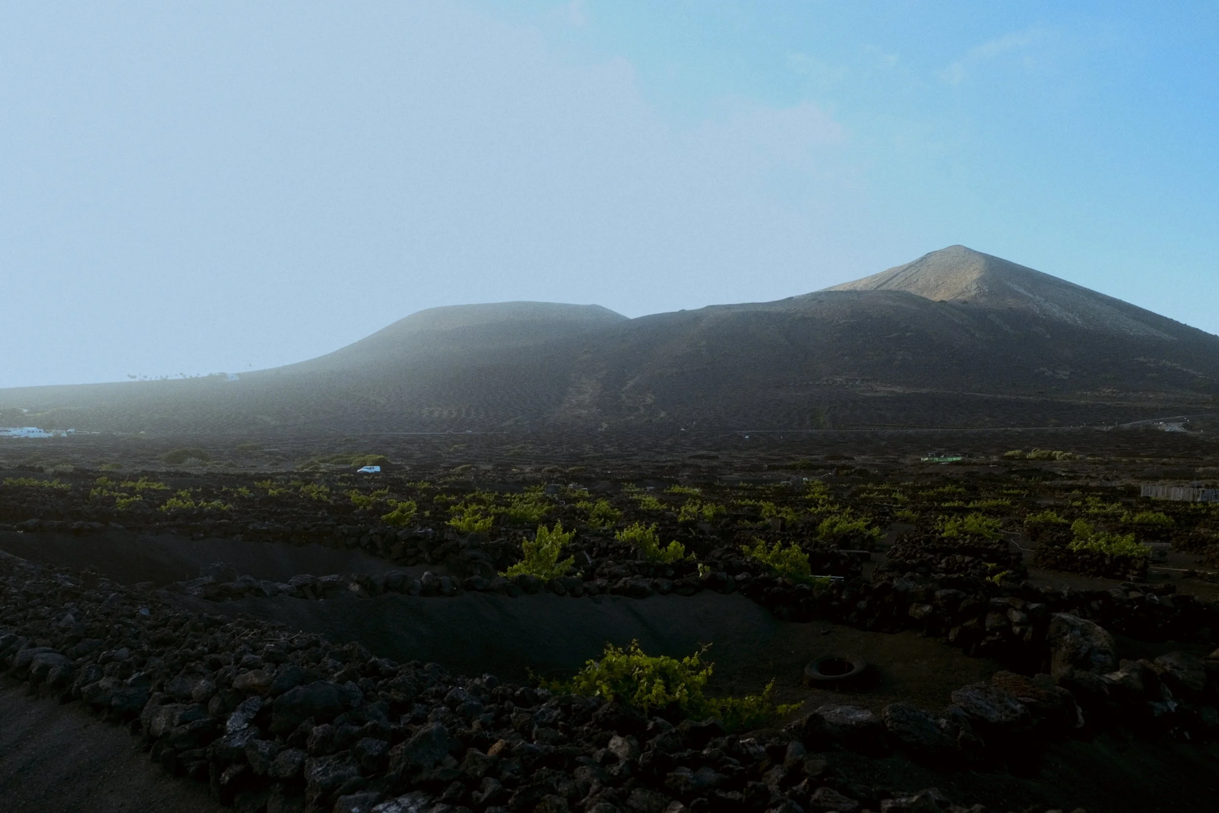 Une montagne volcanique sous un ciel clair, avec une lave noire visible sur ses pentes et quelques végétations au premier plan.