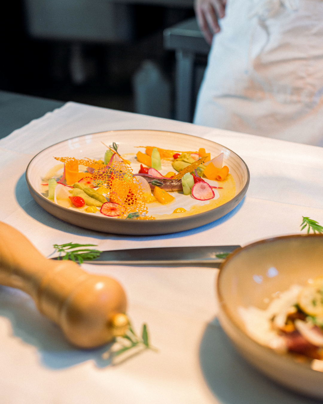 Une assiette de fine cuisine avec des légumes colorés et des décorations artistiques, sur une table blanche, avec un professionnel en arrière-plan.