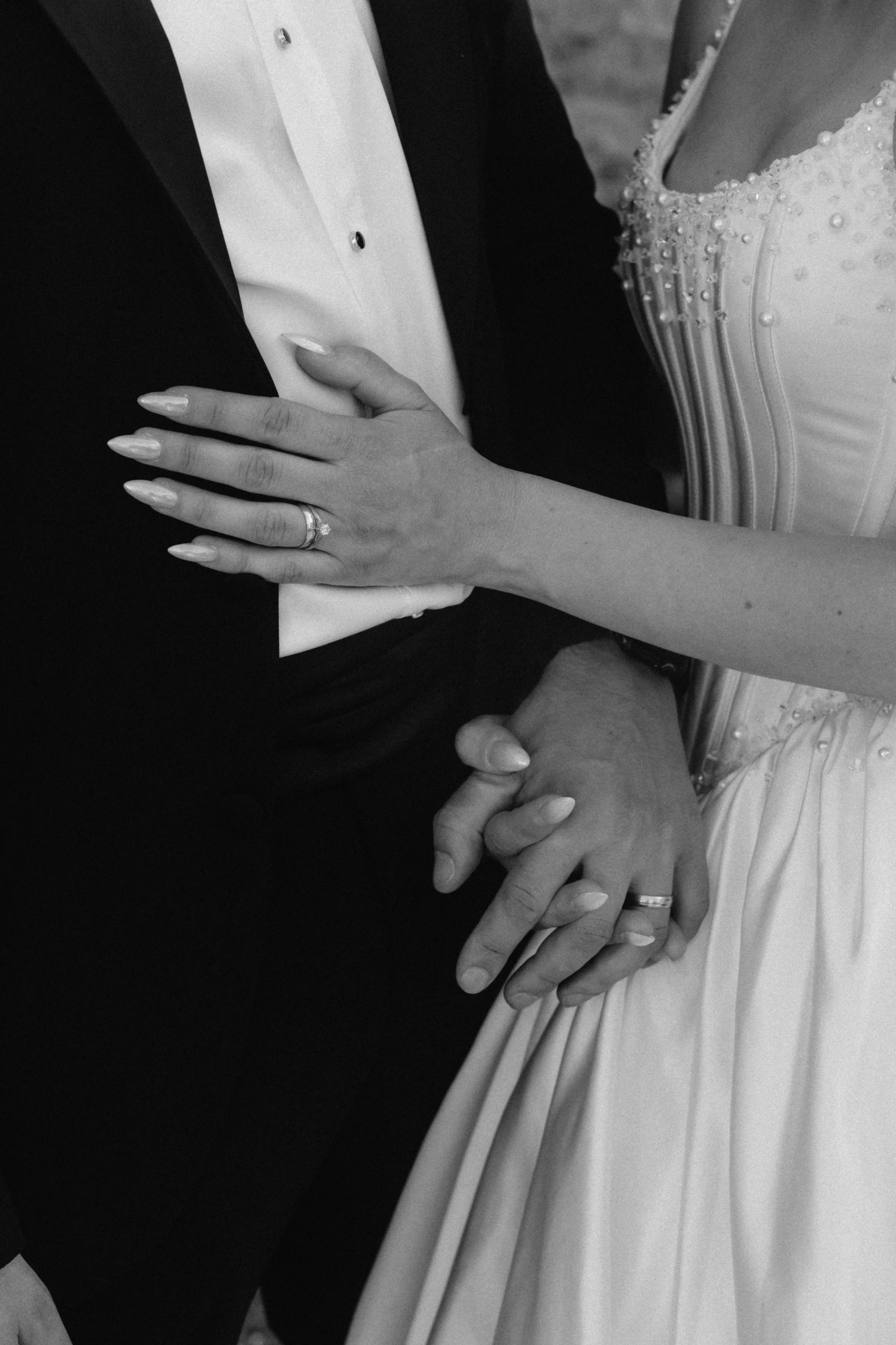 Photo noir et blanc d’un couple lors d’un mariage, montrant les mains entrelacées avec une alliance, et une partie de la robe de la mariée ornée de perles, et la veste du marié visible.
