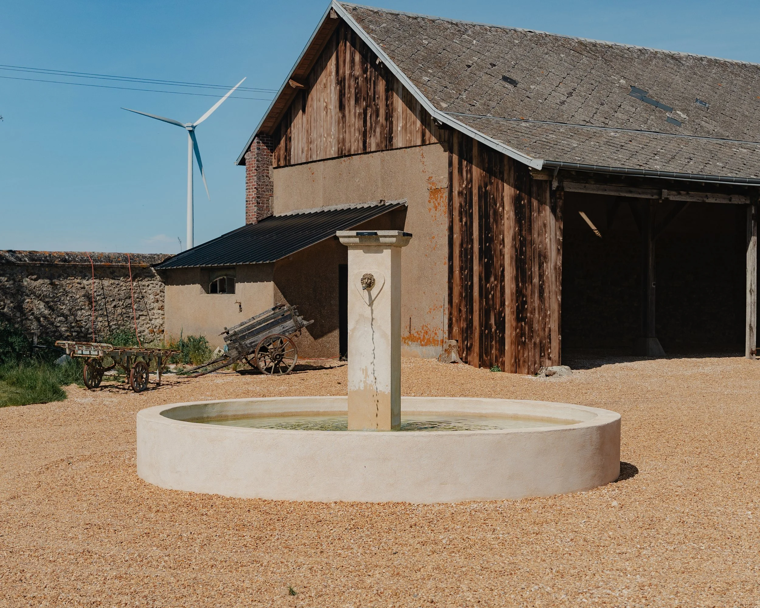 Vieux bâtiment en bois avec un toit en tuiles, fontaine en pierre circulaire au premier plan, dans un espace extérieur avec sol en gravier, ciel bleu en arrière-plan.