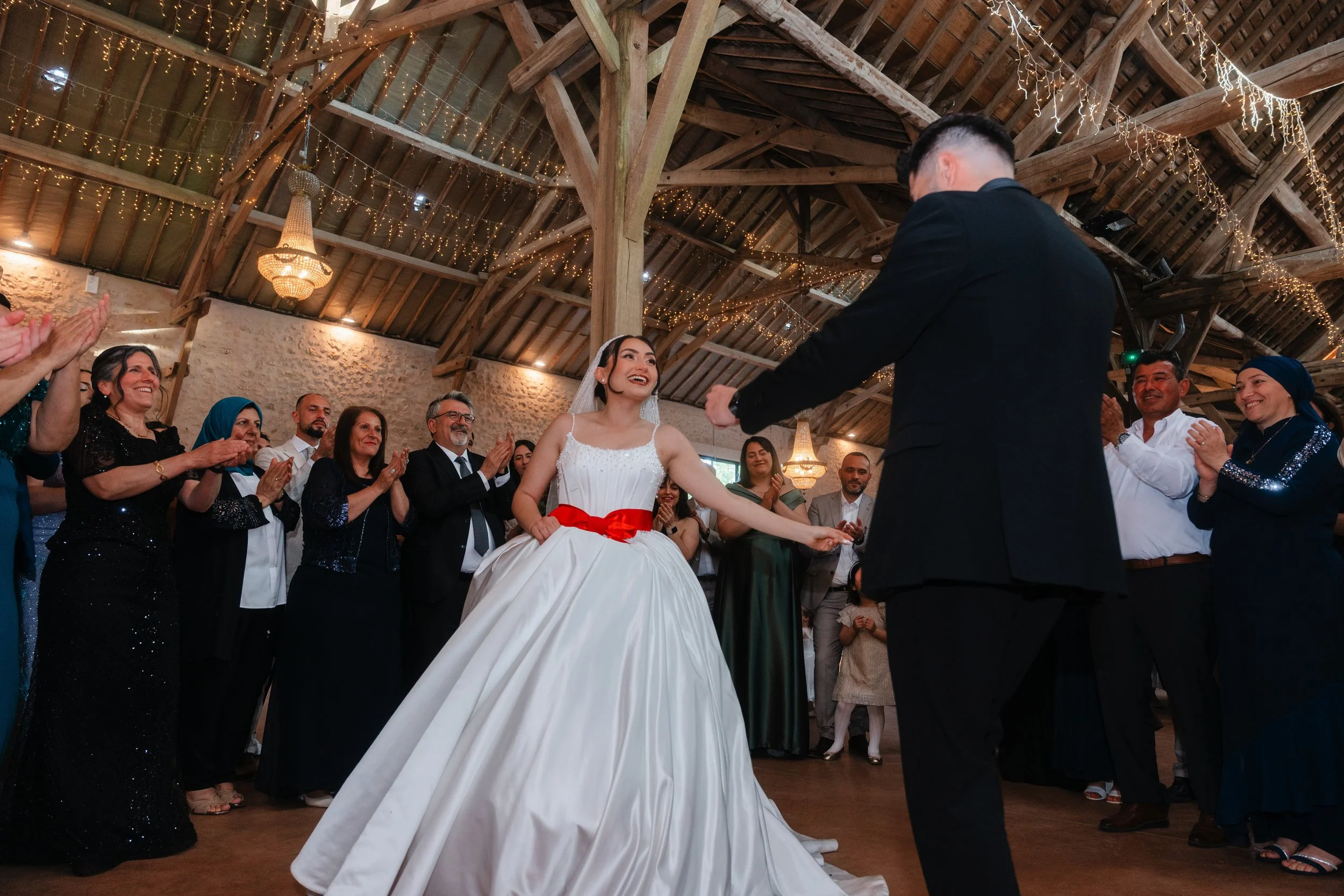 Un mariage avec la mariée en robe blanche assise lors de la cérémonie, entourée de convives souriants dans une salle décorée de guirlandes lumineuses en bois