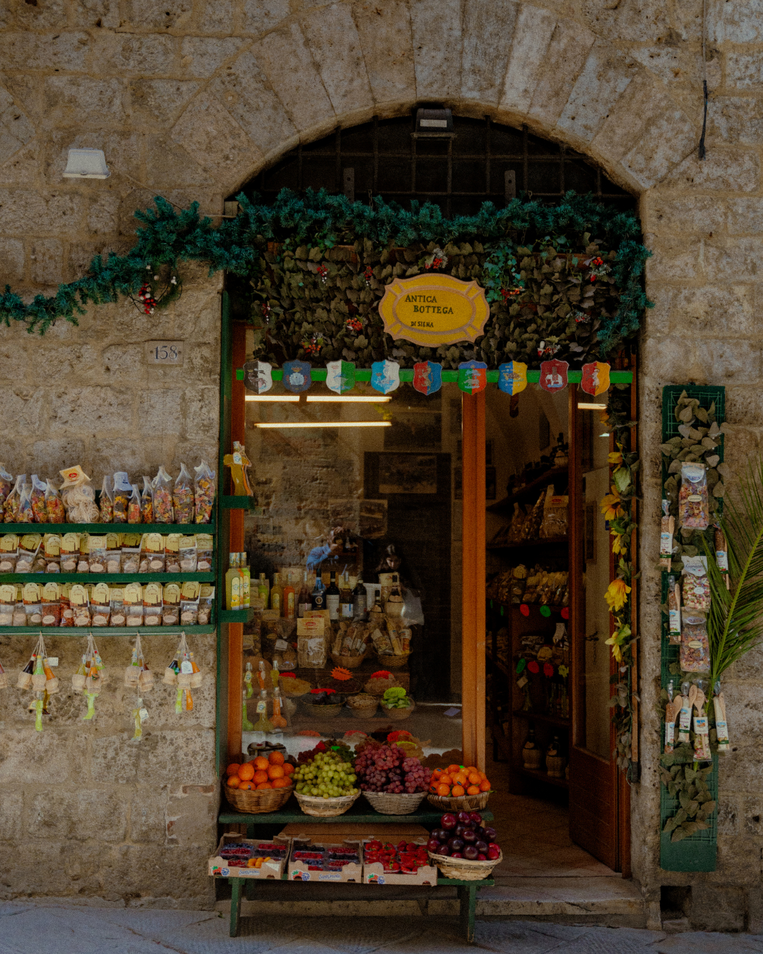 Une boutique de produits locaux avec un décor festif de Noël, présentant des fruits comme des oranges, des raisins, et des pommes, et des sachets de confiseries dans un mur en pierre.