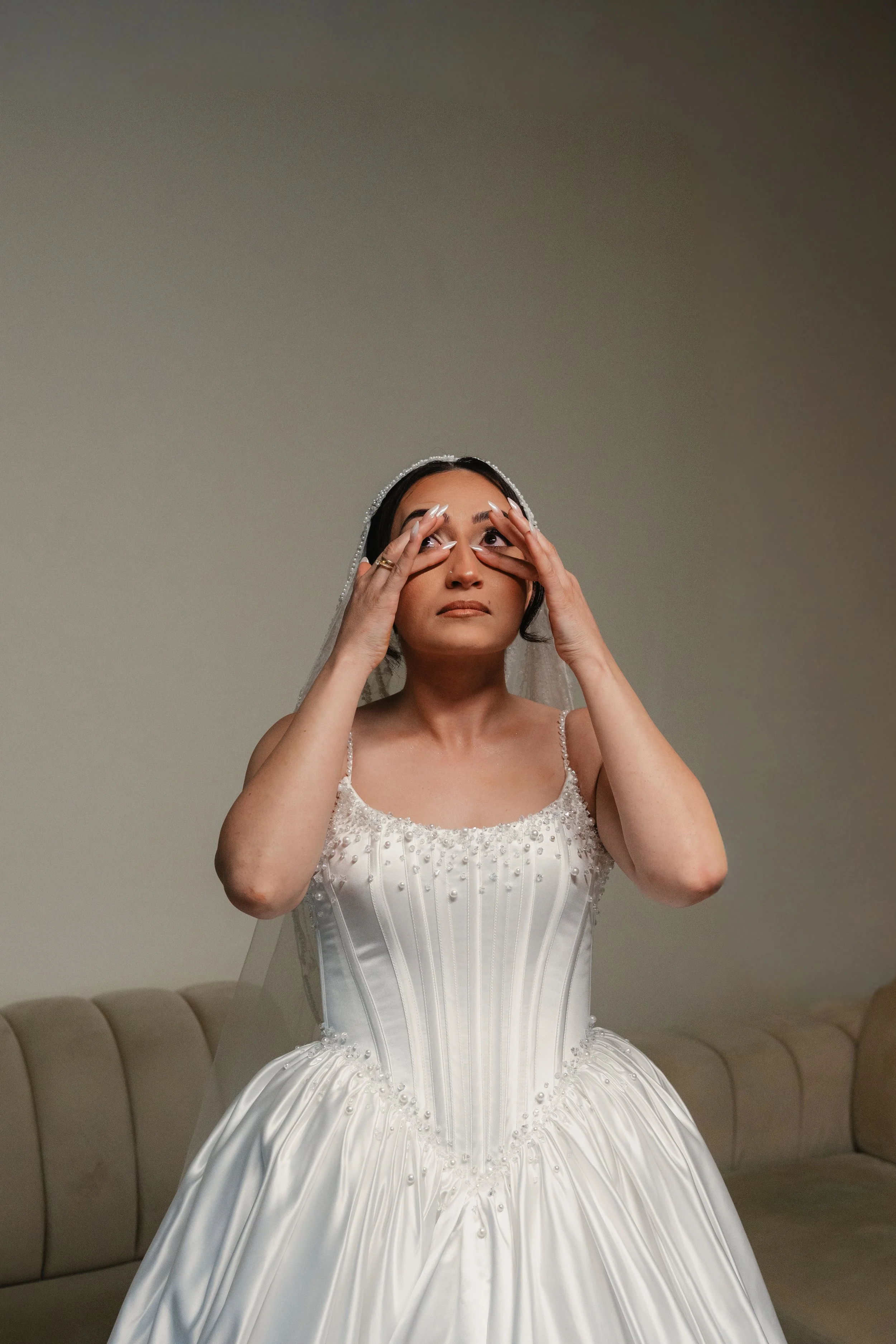 Une femme en robe de mariée blanche, qui ajuste ses yeux avec ses mains, assise sur un canapé beige contre un mur neutre.
