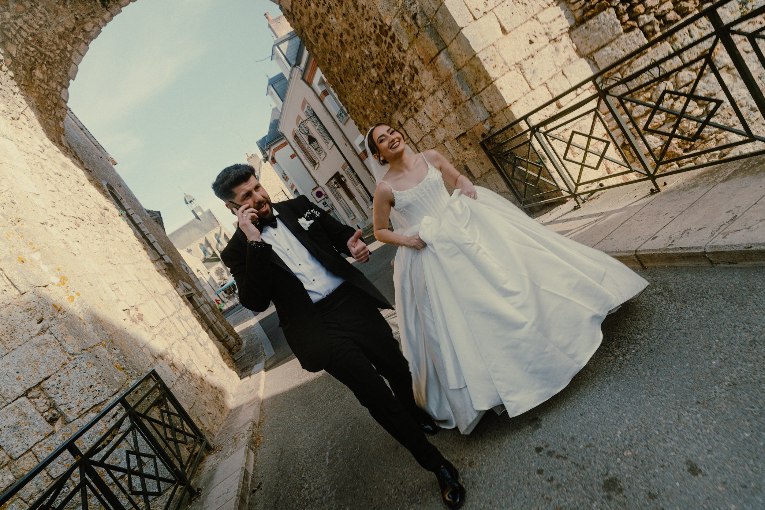 Un couple de mariés souriant et racontant une blague dans une rue pavée, entourée de bâtiments en pierre, en France.