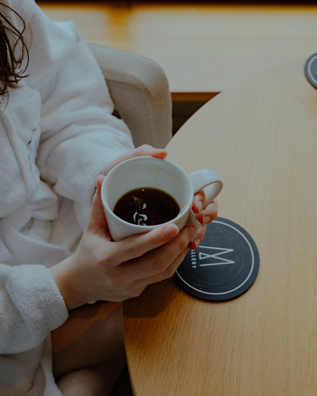Une personne tenant une tasse de café noir dans une main, assise à une table en bois avec un sous-verre noir."