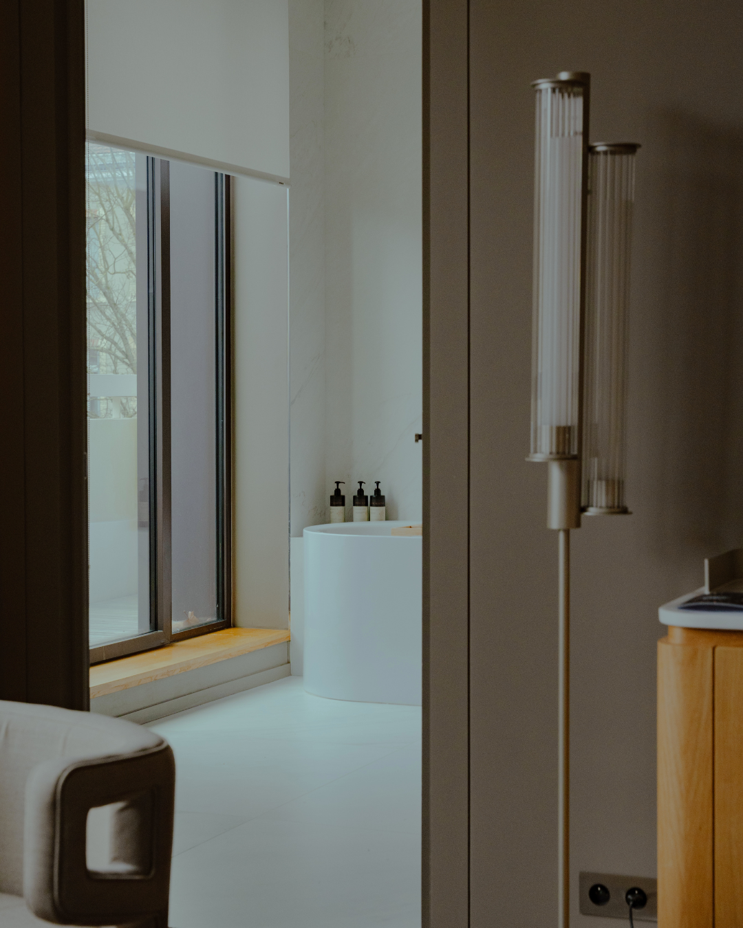 Vue partielle d'une salle de bains avec une baignoire blanche, des bouteilles de produits de bain, une grande fenêtre et un radiateur à rayons.