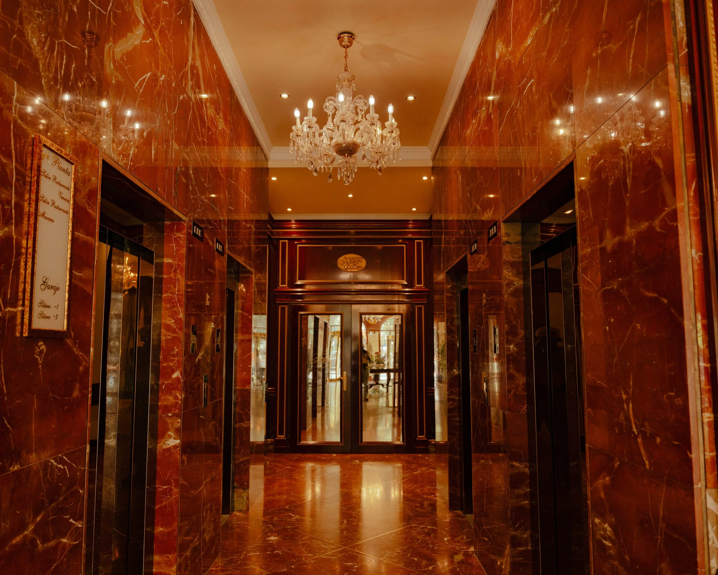 Un hall d'immeuble avec des murs en marbre rouge brillant, un plafond beige avec moulures blanches, un chandelier en cristal suspendu et des éclairages encastrés. On voit également des ascenseurs avec des panneaux à gauche et à droite.