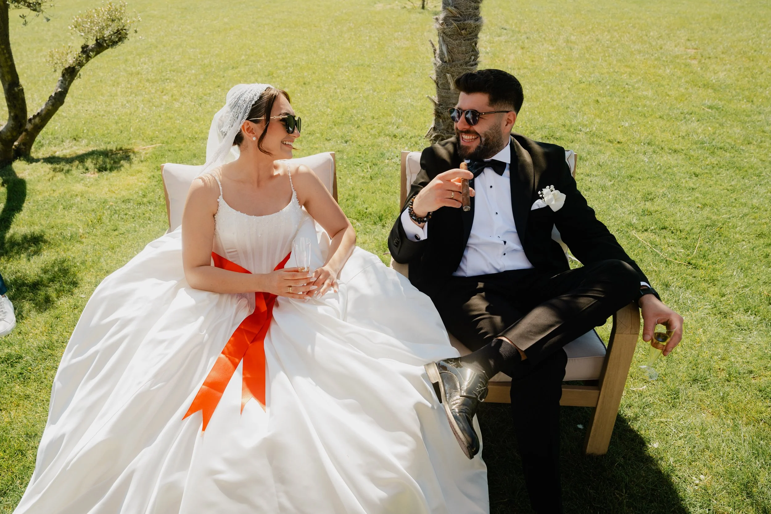 Un couple de mariés, souriant, assis dans une ambiance ensoleillée, avec des lunettes de soleil, une bride blanche et une robe blanche avec un ruban orange, un homme en costume noir avec une chemise blanche, tous deux tenant un cigare.