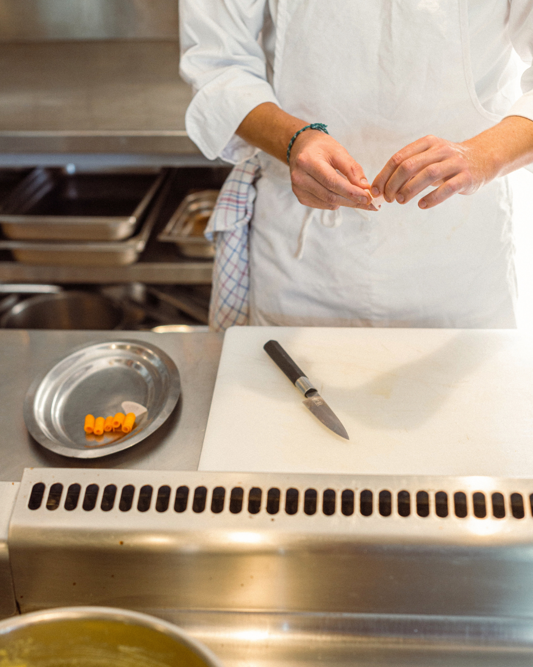 Une personne en chemise blanche prépare des pâtes dans une cuisine professionnelle, avec des raviolis orange sur une assiette en acier inoxydable et un couteau sur une planche à découper.