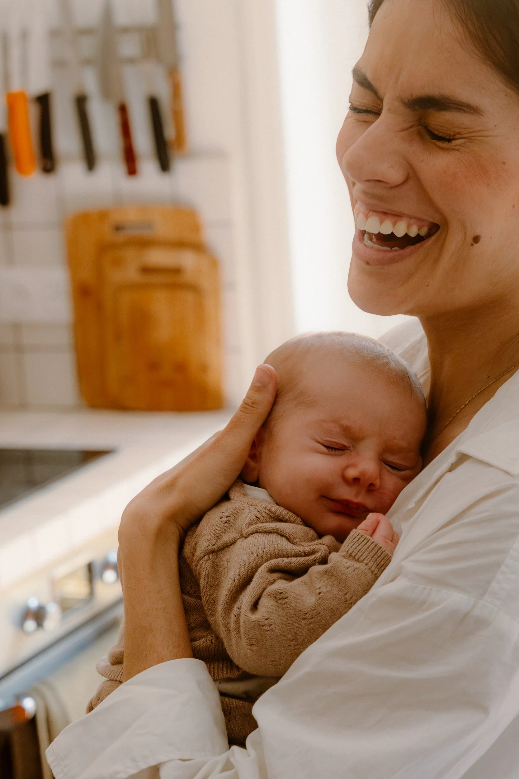 Une femme souriante tenant un bébé endormi dans ses bras, dans un cadre intérieur comme une cuisine.