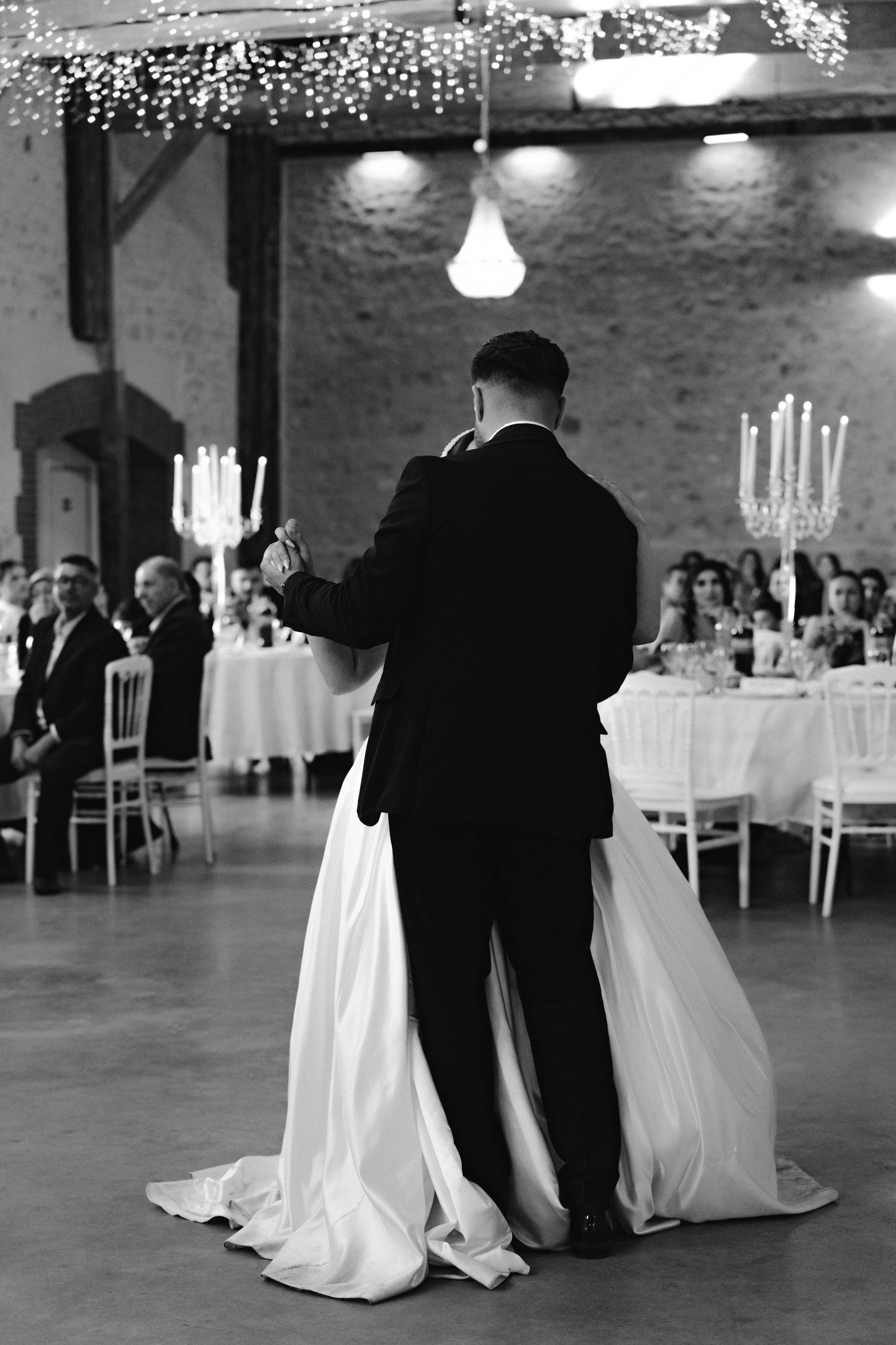 Un couple de mariés dansant lors d'une réception, entourés d'invités assis à des tables décorées, dans une salle éclairée avec des chandeliers et des guirlandes lumineuses.