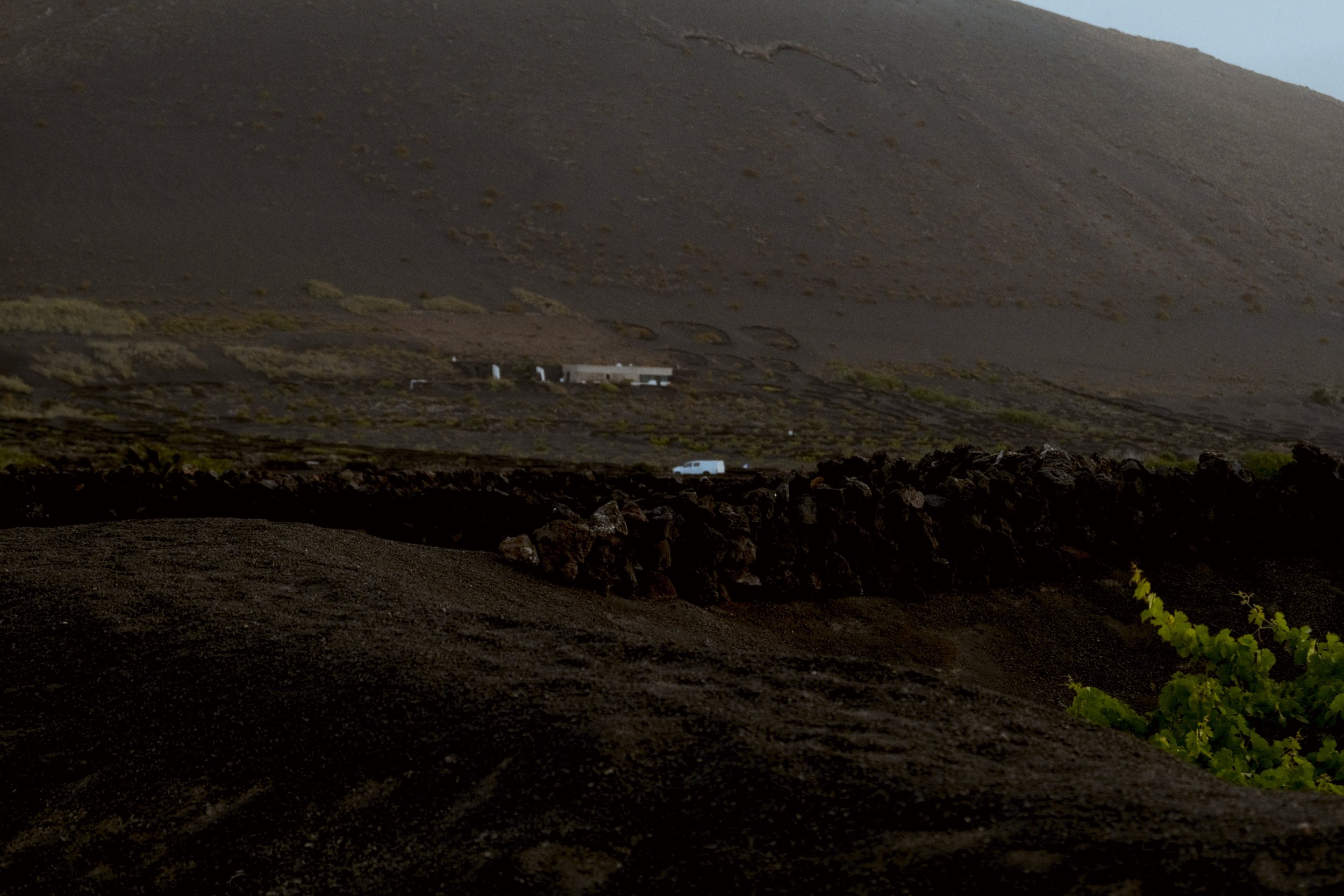 Paysage volcanique avec des bâtiments blancs en contrebas, terrain sombre et monts arides sous un ciel clair.