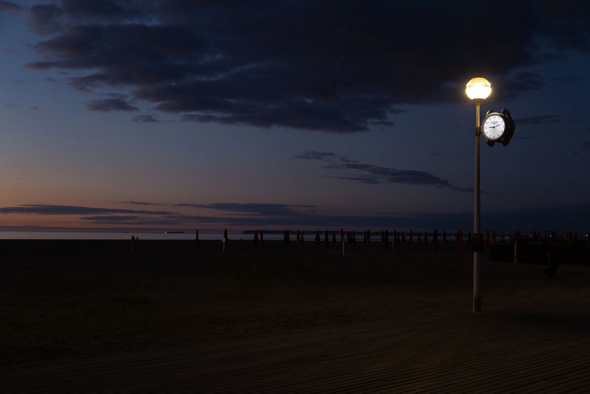 Une horloge sur un poteau se trouve près d'une lanterne allumée lors d'un crépuscule ou d'une aube, au bord d'une plage avec des parasols rouges en arrière-plan.