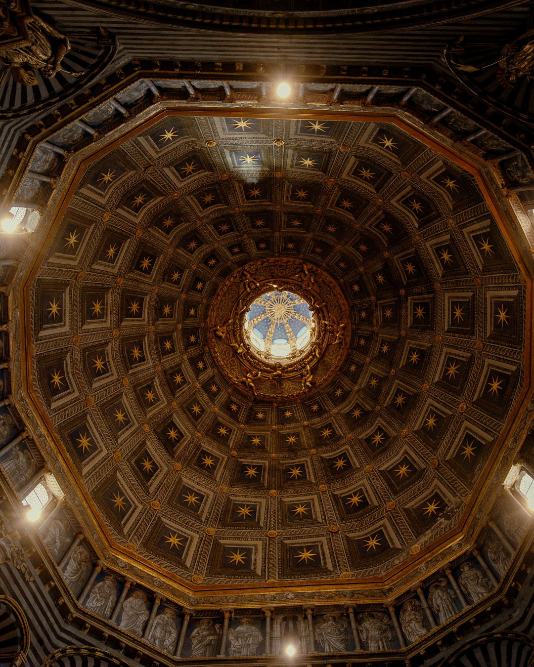 Vue du plafond doré d'une coupole, avec des motifs étoilés et un puits de lumière au centre, décorations sculptées et colonnes ornées entourent la base.