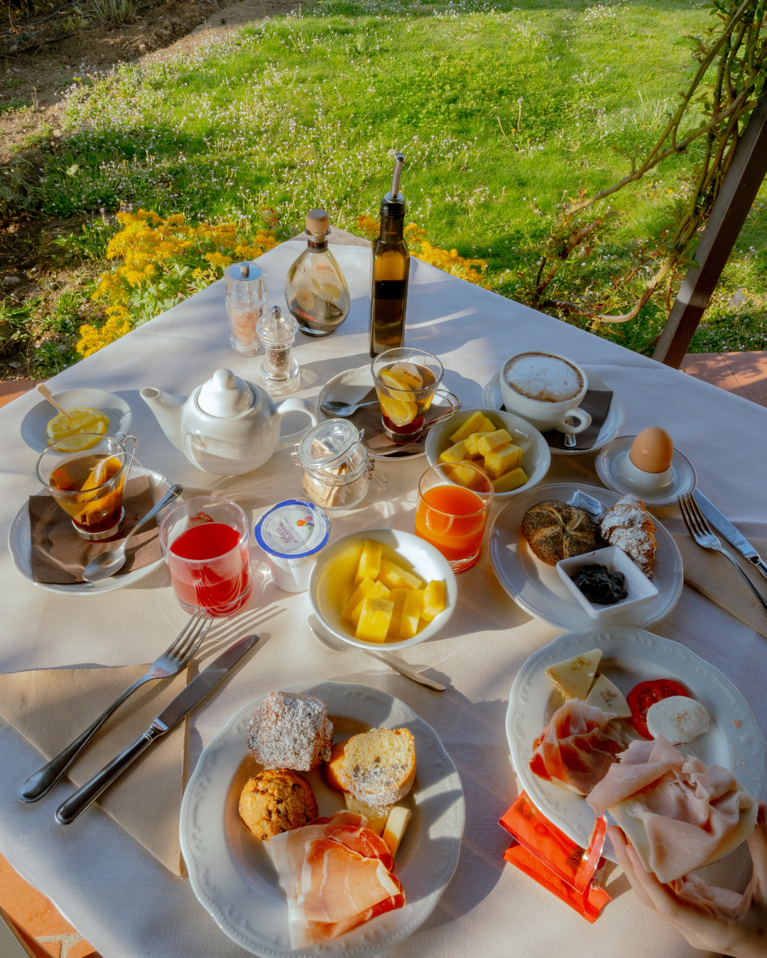 Petit-déjeuner avec œuf, viennoiseries, fruits, jus, thé, et charcuterie sur une table en extérieur.