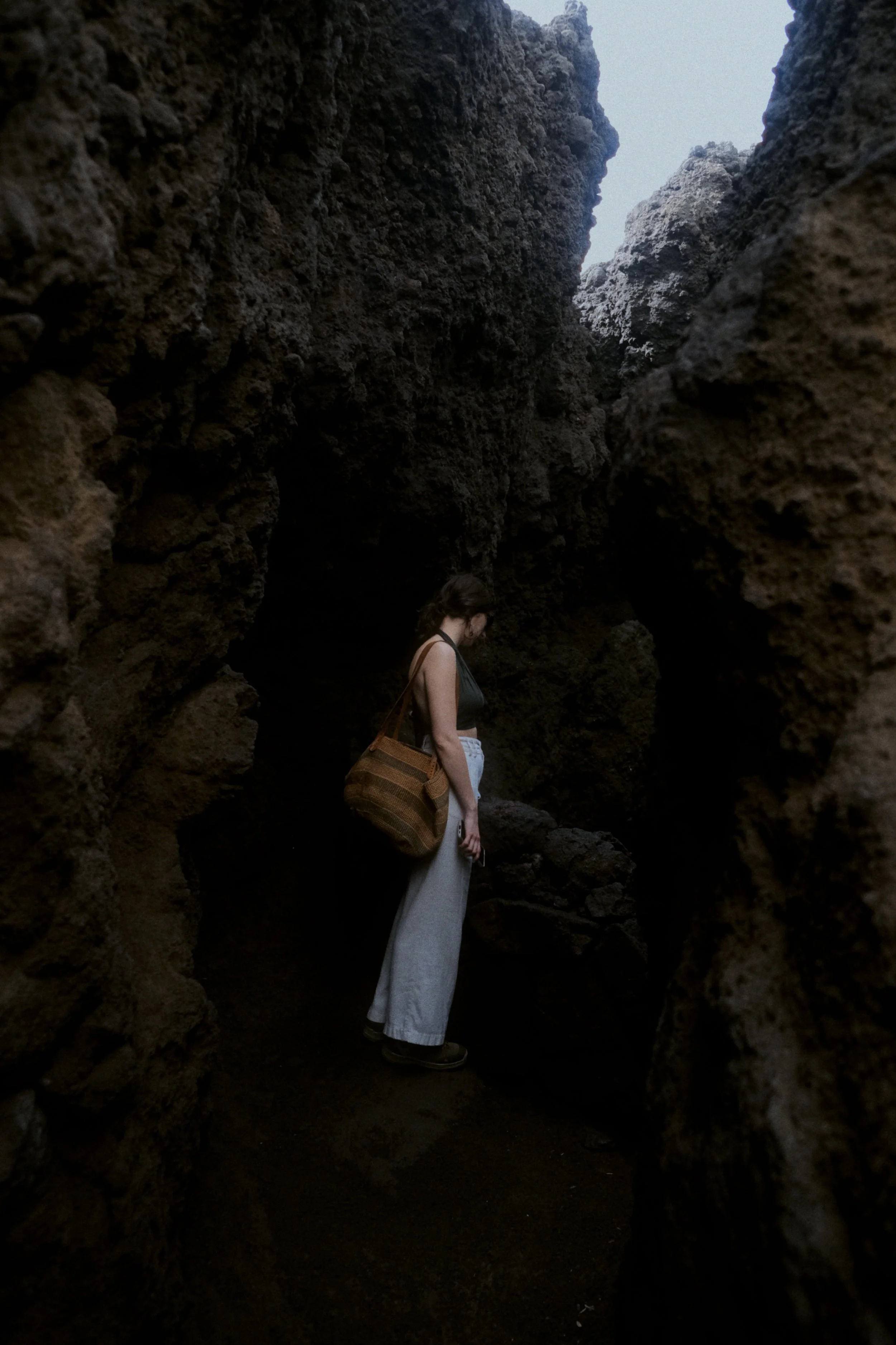 Une femme portant un sac en bandoulière, vêtue d'un haut sans manches et d'une longue jupe, se trouve dans une grotte sombre entourée de rochers volcaniques, regardant vers le bas.