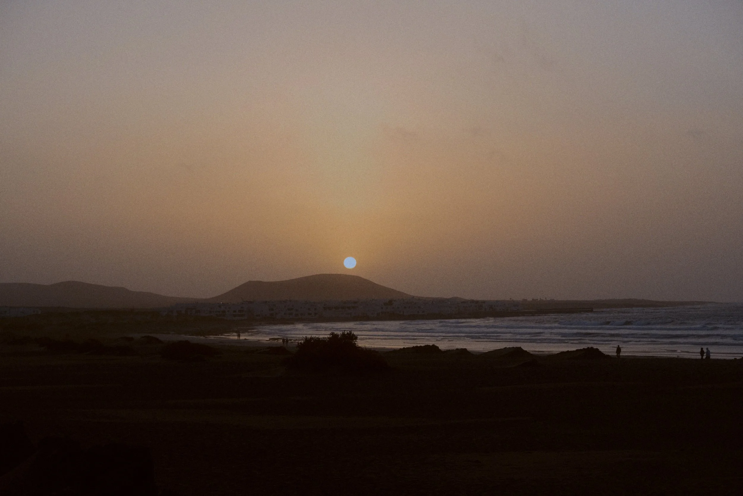 Coucher de soleil sur une plage avec des silhouettes de personnes, des vagues et des montagnes au loin.