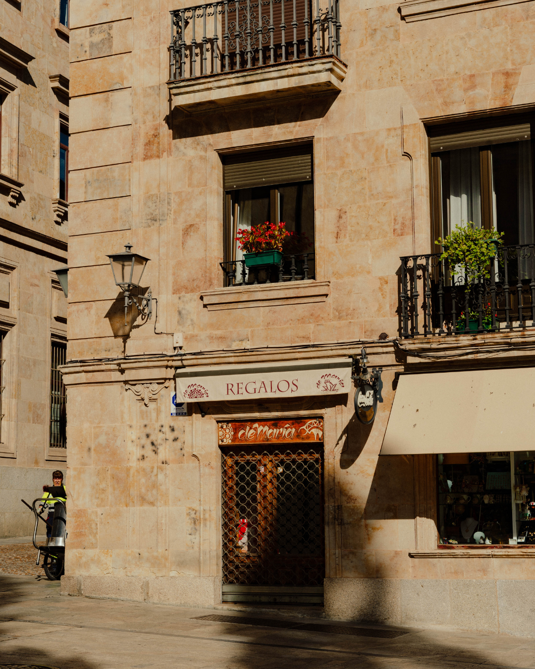Facade d'un bâtiment en pierre avec des balcons en fer forgé, fenêtres avec fleurs, et un magasin avec une grille métallique à la porte.