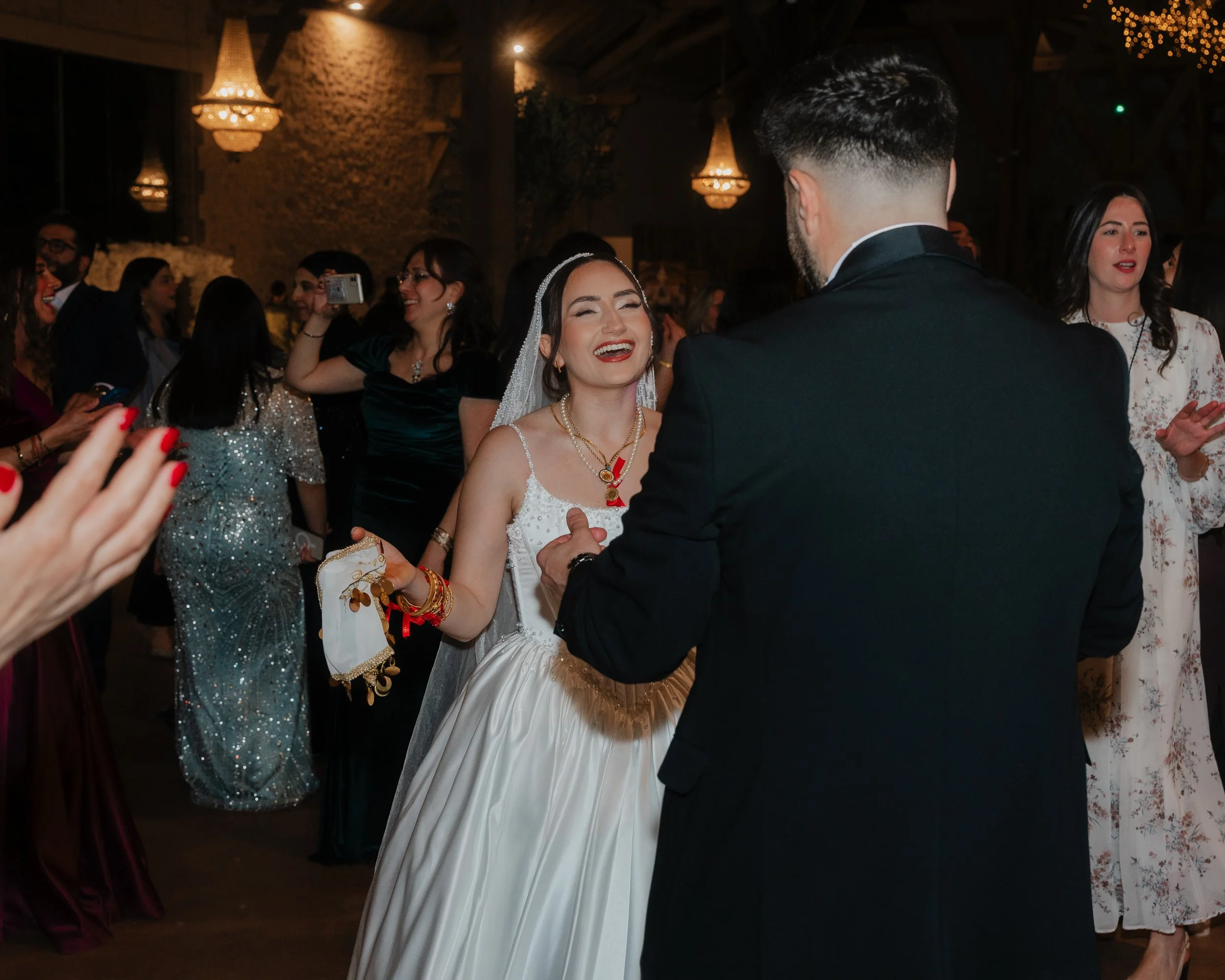 Un couple de mariés danse lors de leur mariage, entourés d'invités qui prennent des photos et applaudissent dans une salle décorée avec des lampes suspendues et des guirlandes lumineuses.